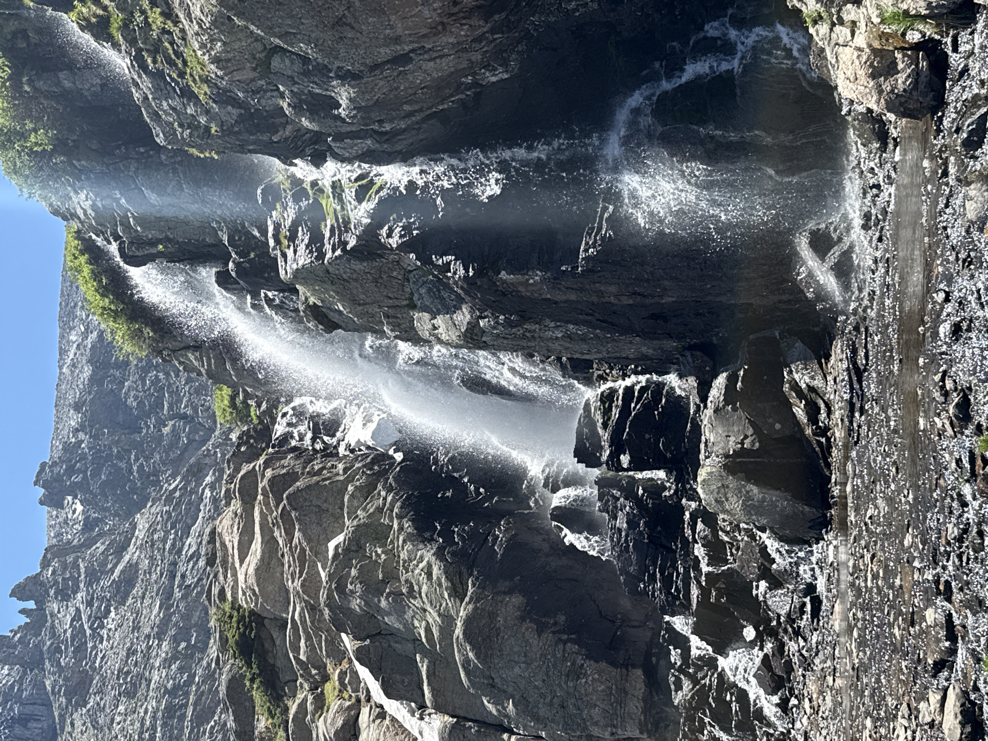 The image shows a waterfall cascading down a rocky mountainside. The water is white and frothy as it falls, and there are many rocks and boulders at the base of the waterfall. Green vegetation is visible on the upper parts of the mountain. The sky is clear and blue. This is a view of the Twin Sisters Peaks in Allenspark, Colorado.