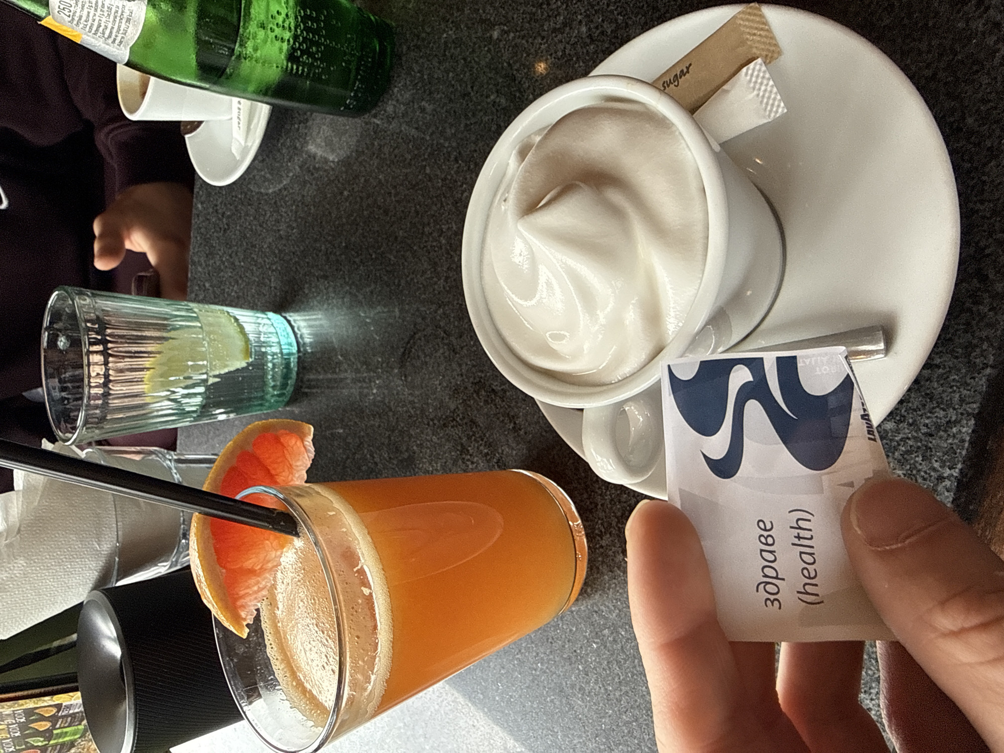 A high-angle shot shows a table setting, likely in a café or restaurant, located in Varna, Bulgaria. An individual (partially visible in the lower left and upper left corners) sits at the table.
On the table is a glass of orange juice with a grapefruit slice, a glass of water, a white teacup filled with white foam (possibly whipped cream or yogurt), a saucer holding sugar packets, and a bottle of green liquid (likely mineral water or soda). A hand holds a packet labeled "здраве (health)", suggesting the packet contains a health supplement or product.
The table surface appears to be dark gray or black and is textured. Overall, the setting suggests a relaxed atmosphere of someone enjoying refreshments in a café or restaurant, presumably in Varna, Bulgaria.