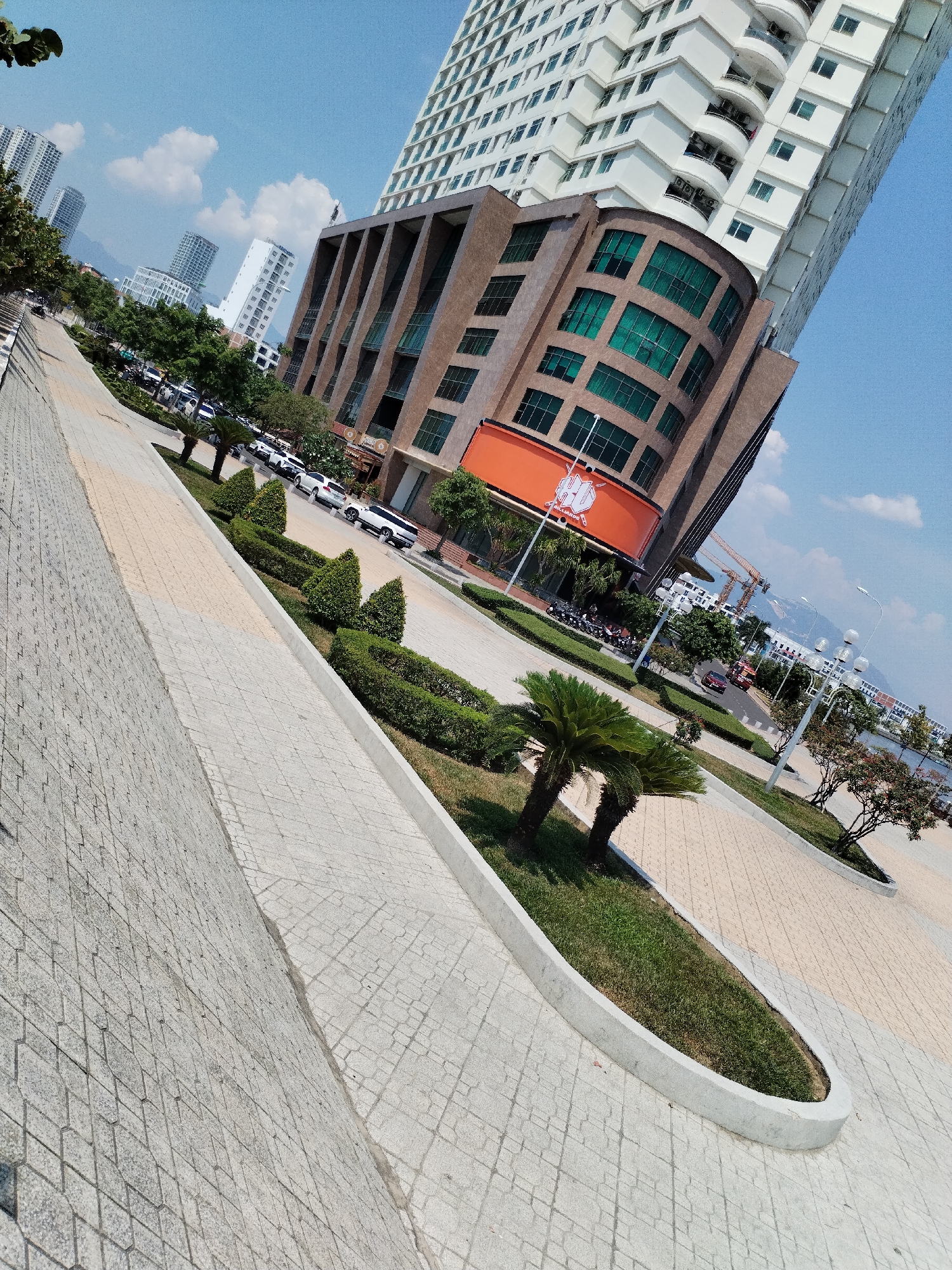 The image depicts a street scene in Nha Trang Ward, Vietnam, captured from a high-angle perspective. A modern high-rise building, with a distinctive curved architectural element featuring green-tinted windows above an orange signage, is prominently displayed. A taller, white tower rises behind this structure. The street view shows a well-maintained paved walkway alongside a road with parked and moving vehicles. Landscaped elements, including manicured bushes and small palm trees, are strategically placed along the walkway. In the background, distant buildings, possibly hotels or residential apartments, are visible. The sky is partly cloudy, suggesting a sunny day. The image conveys a sense of urban planning and modern architecture within a tropical or coastal city setting. No people are distinctly visible in the image. Construction cranes are visible in the far background.