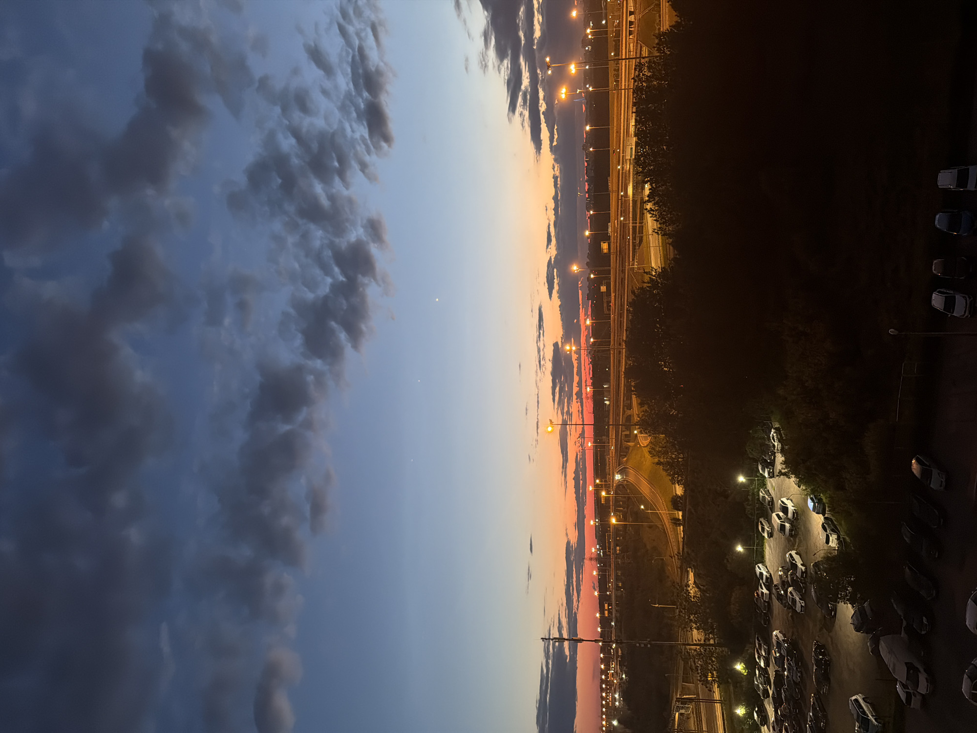 This is an eye-level, high-angle medium shot of a city skyline at dusk. In the foreground, there is a dark, silhouetted line of trees. Behind the trees, a parking lot is illuminated by streetlights, filled with numerous parked cars. Beyond the parking lot, a multi-lane highway curves through the landscape, with more streetlights lining its paths. In the distance, the city skyline is visible, with buildings and more illuminated roadways. The sky transitions from a deep blue at the top to a fiery orange and red near the horizon, with scattered clouds. Two bright celestial bodies are visible in the upper sky.