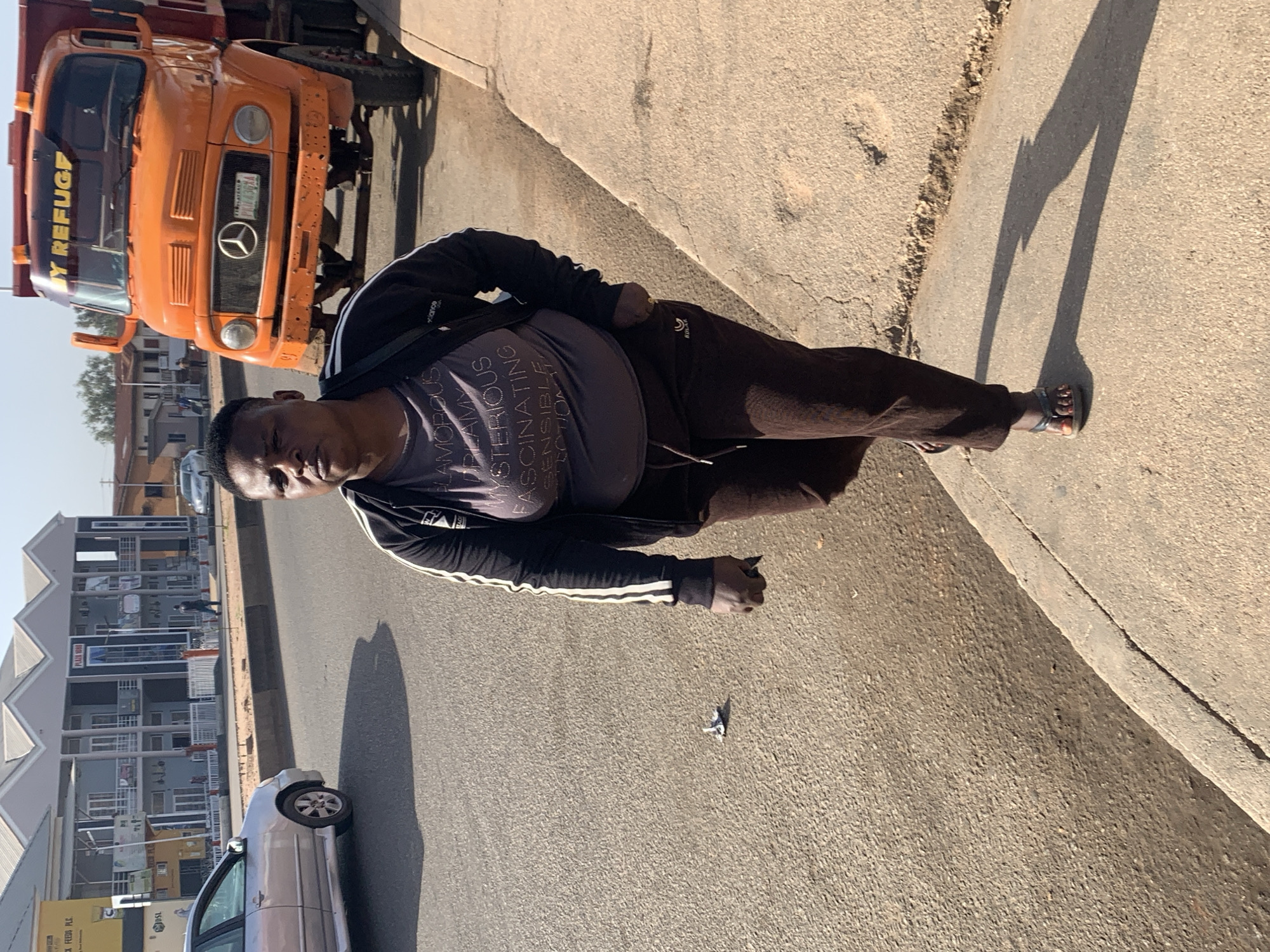 A medium-shot depicts a person standing on an asphalt road next to a concrete curb. The individual is facing left, wearing a dark-colored t-shirt with light-colored text, a dark jacket with white stripes, and dark pants. They are also wearing sandals. An orange truck with the text "Y REFUGF" visible on the front is parked on the left side of the frame. Behind the person and the truck, a silver car is visible on the road. In the background, several buildings are lined up, and signage reads "PLAJA 1080" and "BLACK FEEDS PLC". A small piece of litter lies on the road near the individual. Given the signage "PLAJA 1080", "BLACK FEEDS PLC", and the presence of the orange truck, the location is likely in Jos, Nigeria.
