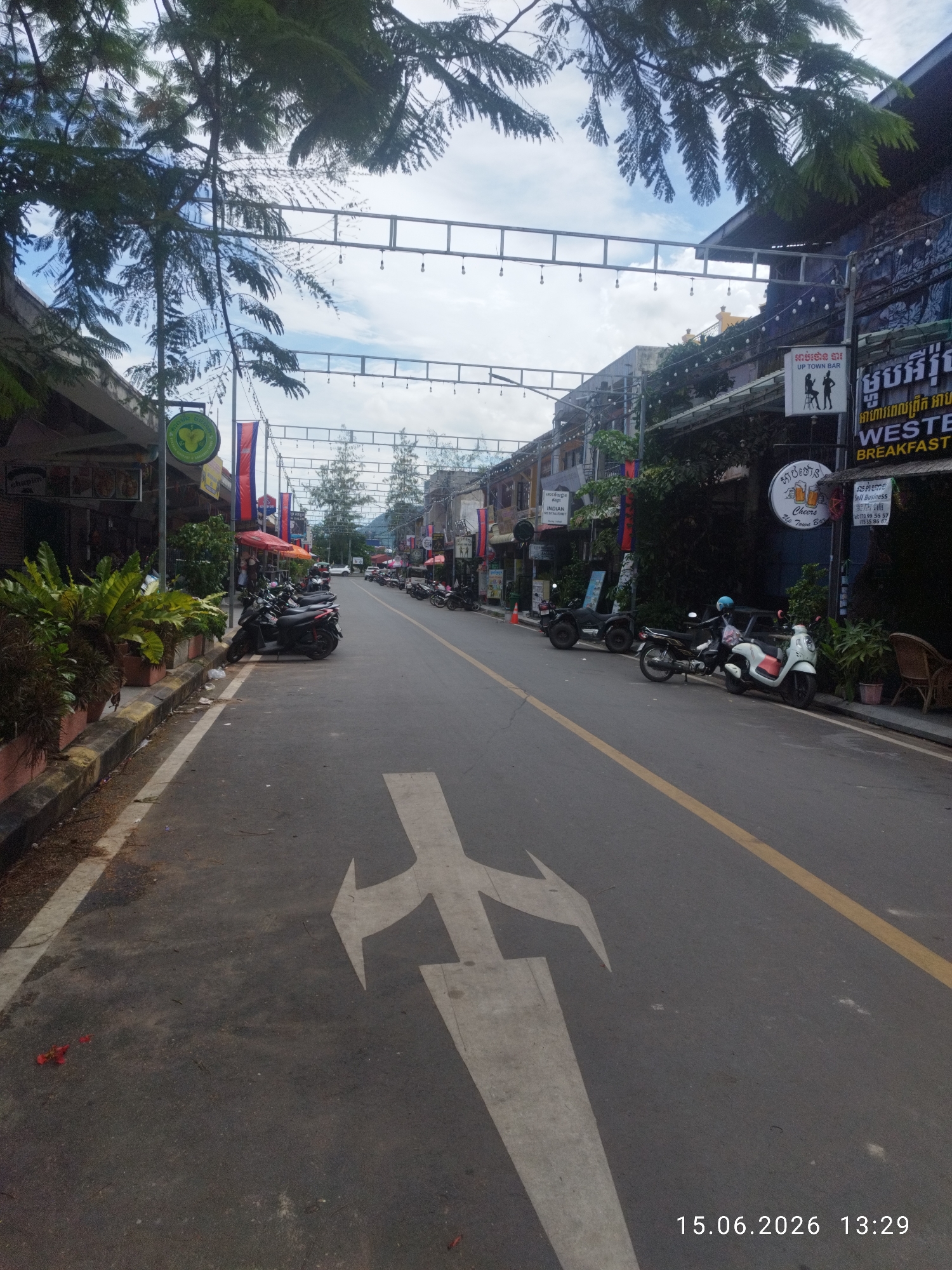The image shows a street scene in Kampot, Cambodia. The street is paved and lined with buildings on both sides. There are several motorcycles parked along the side of the road, and some businesses with signage in Khmer script are visible. Trees and foliage are present, creating a somewhat tropical atmosphere. The sky is cloudy. A large white arrow marking indicating a turn is painted on the road in the foreground. The timestamp "15.06.2026 13:29" is visible in the lower right corner of the image.