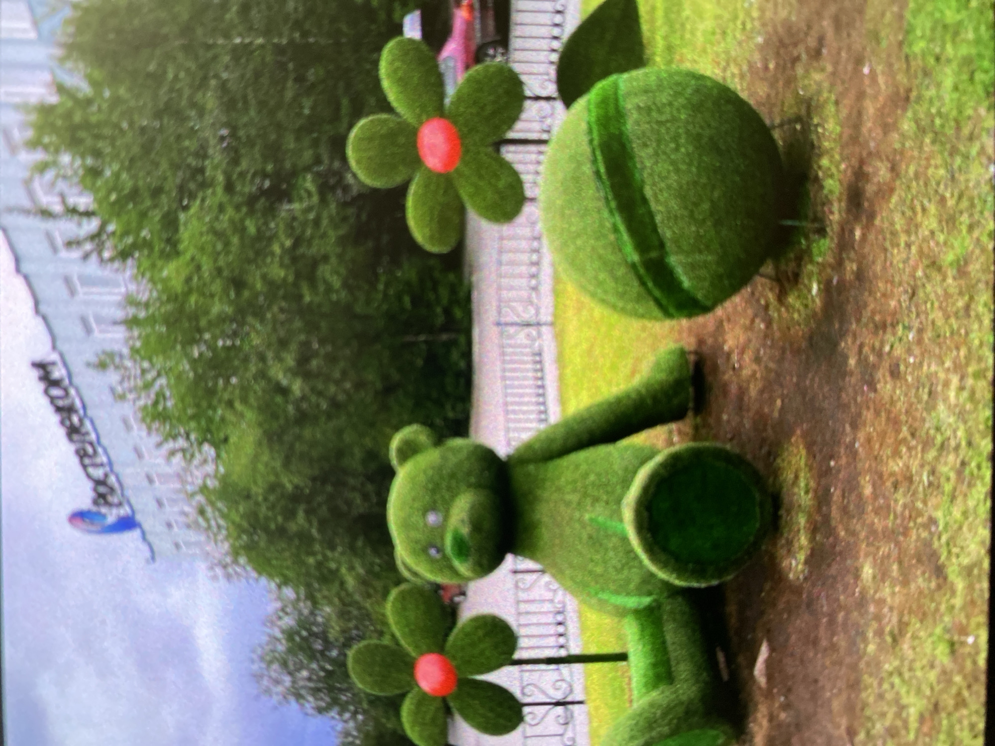 The image shows a street scene in Saint Petersburg, Russia featuring topiary sculptures of a teddy bear, flowers, and a ball. The topiary is made of green material, likely artificial grass or moss. The flowers have red centers. The sculptures are situated on a grassy patch near a fence. In the background, there's a building with signage that includes the word "РОСТЕЛЕКОМ" (Rostelecom), a Russian telecommunications company. The background shows trees and possibly other buildings, suggesting a park or urban environment.