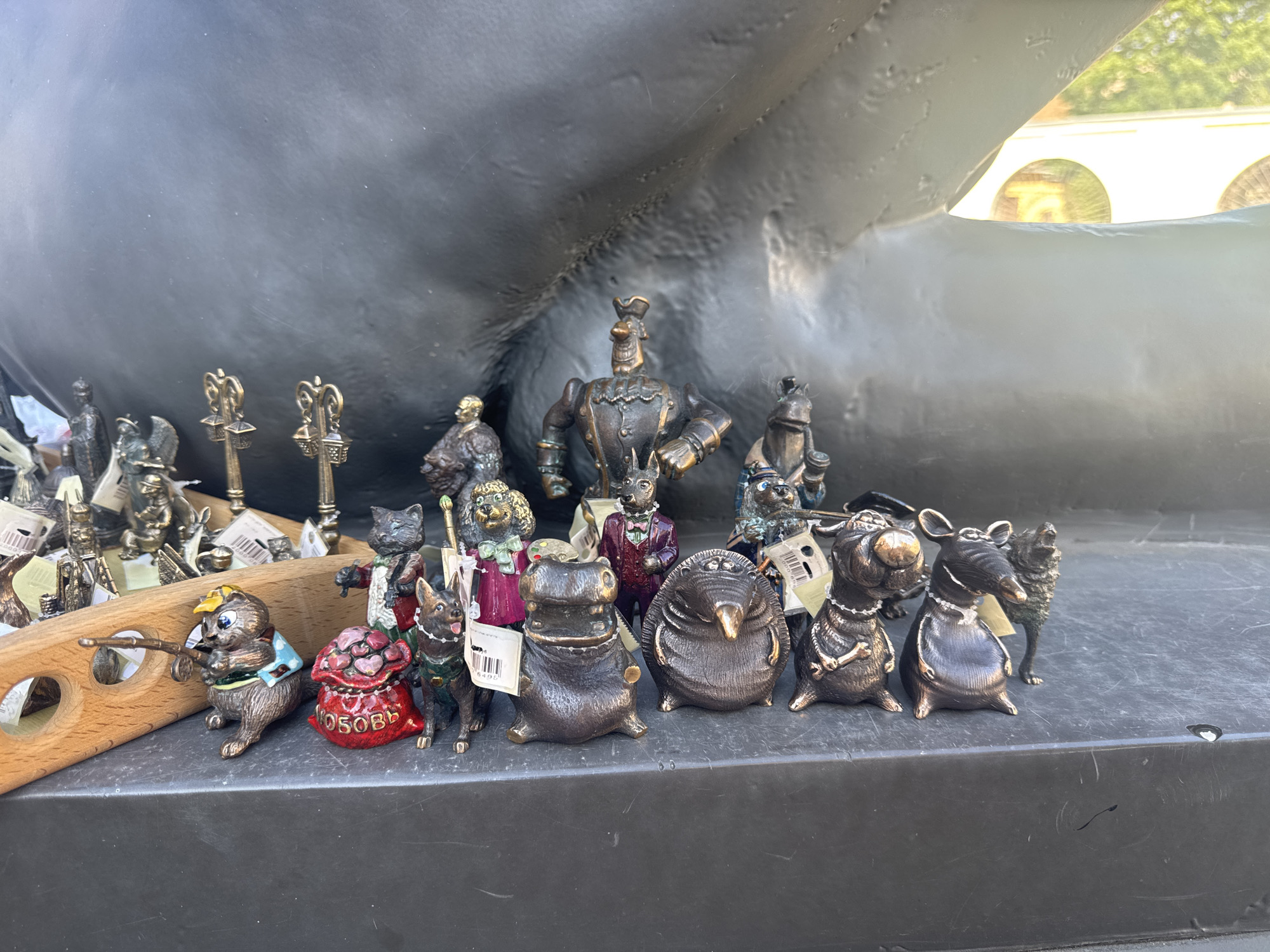 The image shows a collection of small bronze figurines arranged on a dark surface, possibly a ledge or a display. The figurines depict various animals and anthropomorphic characters. In the foreground, from left to right, there is a small animal figurine, possibly a hamster or squirrel, wearing a blue shirt and yellow hat, holding a fishing rod. Next to it is a red bag filled with heart shapes, with "ОСОБОВ" written on it. Following this is a figurine of a dog wearing a green vest, and then a stylized hippopotamus. To the right of the hippopotamus is a round, spiky figurine, resembling a hedgehog or a porcupine. Further to the right are two dog figurines, one with a bone-shaped necklace, and a wolf-like figurine. In the background, there are more figurines of animals and human-like characters, including what appears to be a robot-like figure in the center. The figurines are detailed and appear to be made of aged bronze, with a patina. Behind the figurines is a large, dark, curved object, possibly part of a sculpture or a large building. Sunlight is visible from the upper right, casting a soft light on the scene.
This scene is likely located in Saint Petersburg, Russia, given the typical presence of such souvenir stalls and small sculptures in tourist areas. The style of the figurines, particularly those with a folk art or whimsical theme, is also common in Russian craft markets and street vendor displays. The backdrop, with its dark, monumental form and hint of classical architecture visible through a gap, could suggest a location near a significant landmark or a museum complex in the city.