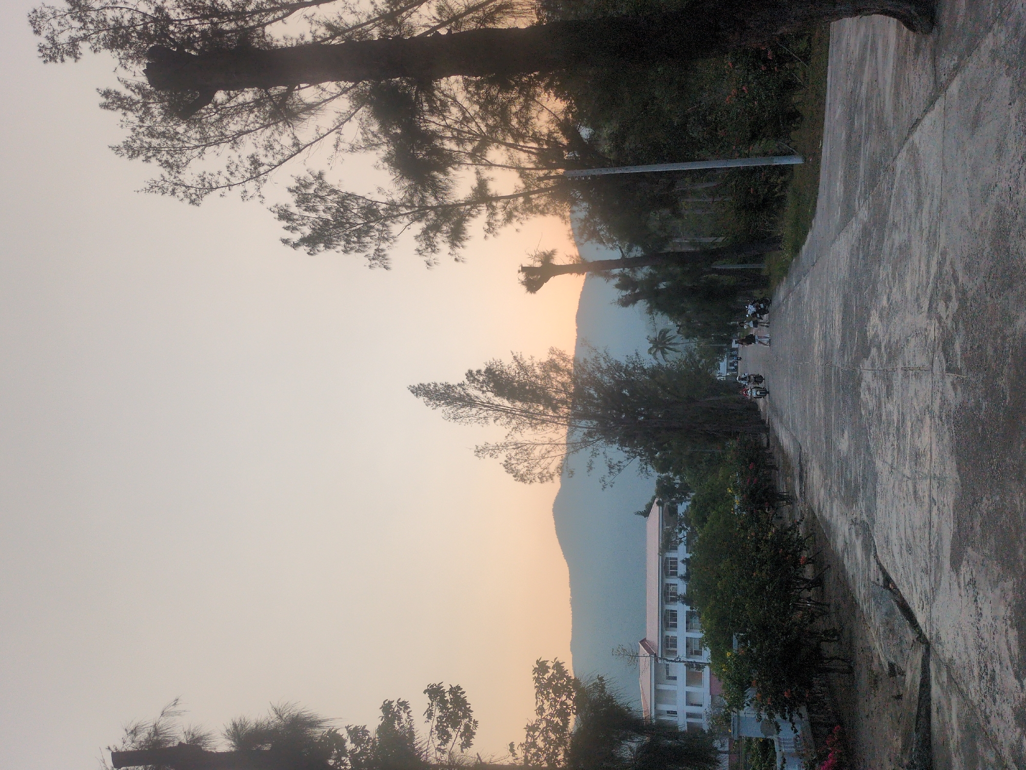 The image shows a street scene in South Quy Nhon Ward, Vietnam, at sunset. In the foreground is a concrete sidewalk with a large crack. To the left of the sidewalk is a line of trees and bushes. Further in the distance is a multi-story white building with a red roof. Behind the building is a mountain range silhouetted against the setting sun. On the road, small groups of people and scooters can be seen. A tall street light can be seen between the trees. The sky is a pale yellow-orange near the horizon, fading to a hazy white. Silhouette of a tree is in the left upper corner.