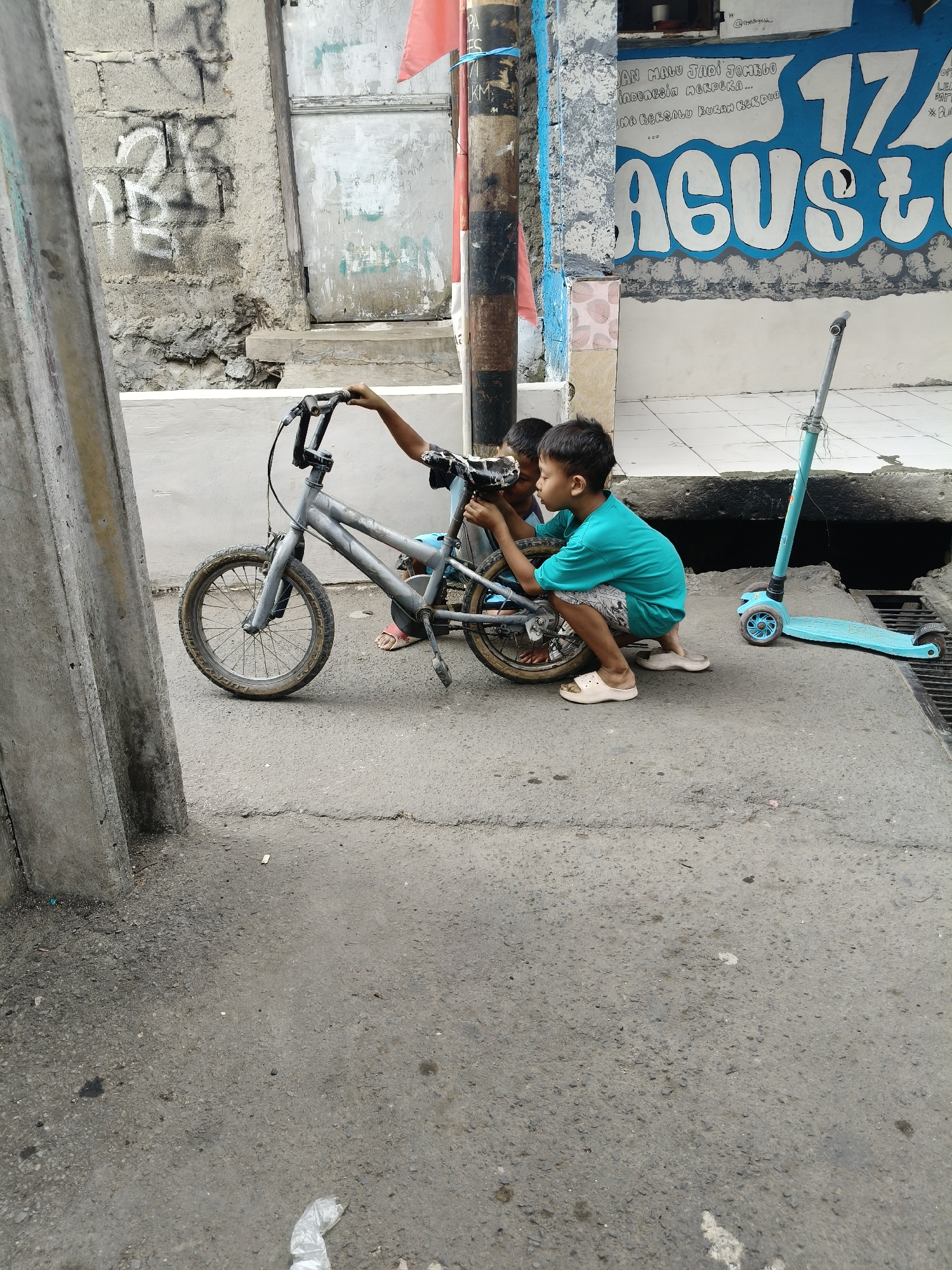 Two young boys are working on a bicycle in front of a building with blue and white graffiti that reads "17 AGUSTUS". The boy on the right is wearing a teal t-shirt and patterned shorts, crouching to adjust the bike. The boy on the left is wearing a black t-shirt and is partially obscured. A teal scooter is parked to the right of the boys. The ground is paved with asphalt, and there are some cracks and debris. The building in the background has a rough, textured wall and a metallic door. The scene appears to be in an outdoor urban setting. The graffiti suggests the image was taken around Indonesian Independence Day. This location is likely an alley or a less maintained area in South Tangerang, Indonesia.