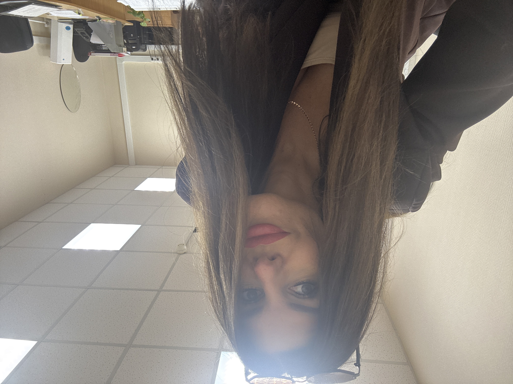 The media file is a selfie of a person with long, brown hair. The person is wearing a light-colored t-shirt and a dark-colored jacket. Their face is partially obscured by their hair. They are wearing red lipstick and have dark eye makeup. They are also wearing glasses. The background appears to be an office or indoor setting with a tiled ceiling and fluorescent lighting. There is a desk in the background with some papers and equipment. The image is taken from a low angle, and the person appears to be looking up at the camera. The lighting suggests it is daytime. No visible text is present.