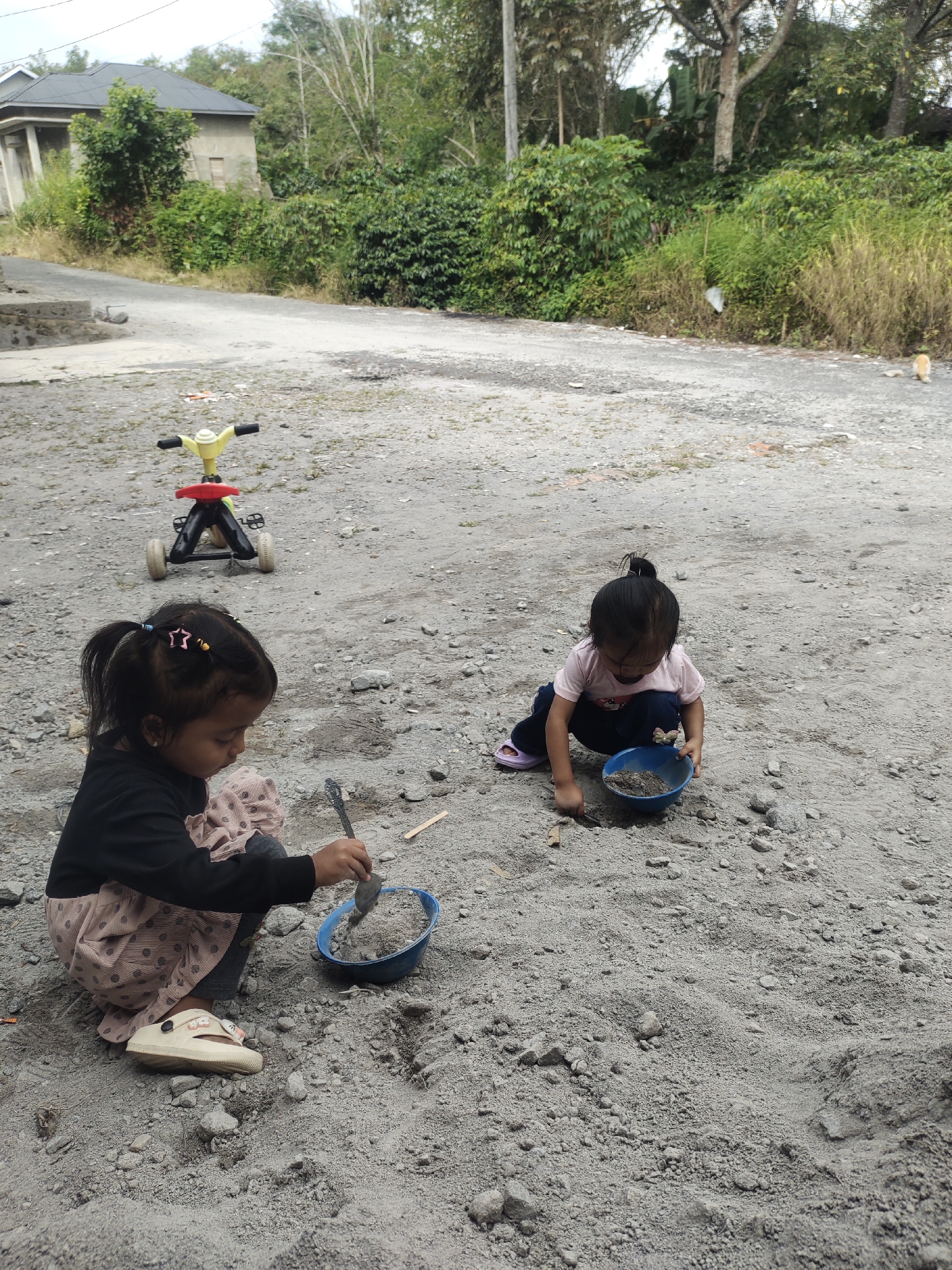 Two young girls are playing in a sandy, outdoor area near a road in Cintadamai, Indonesia. The girl on the left, wearing a black long-sleeved shirt and a patterned pink skirt, is kneeling and using a small shovel to transfer sand into a blue bowl. She has her hair in two ponytails with star-shaped clips. The girl on the right, in a pink t-shirt and dark pants, is also kneeling and appears to be playing with sand in a blue bowl. Behind them, a yellow and red toy tricycle is parked on the gravel. In the background, a simple house with a dark roof is visible, surrounded by lush green trees and vegetation. The ground is a mix of dirt and small stones.