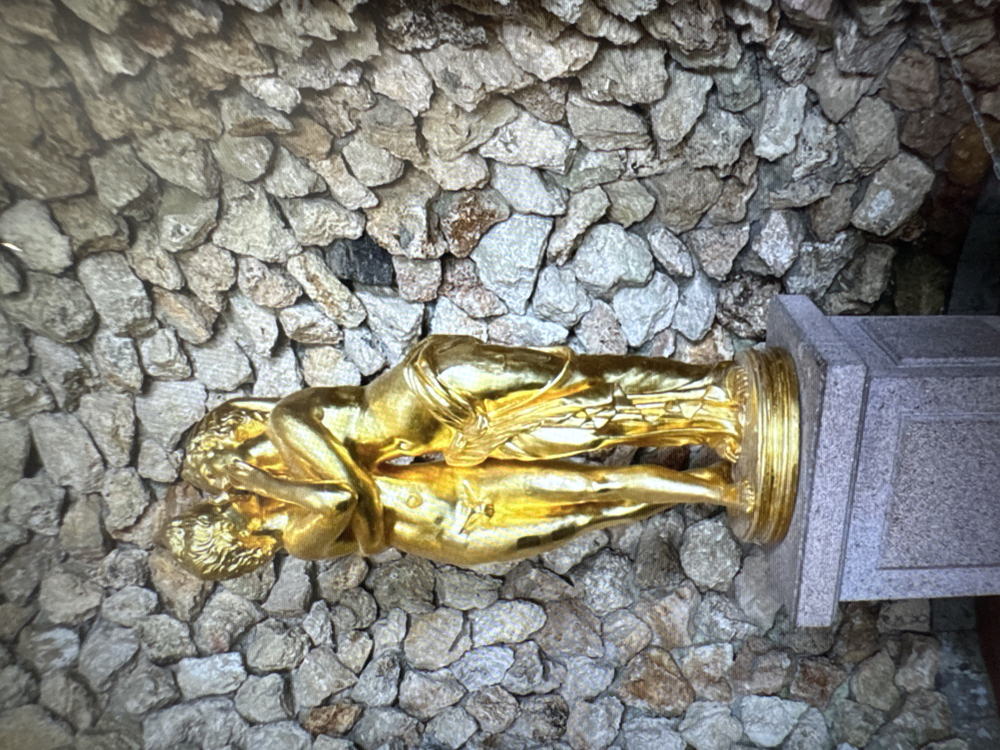 The image presents a golden statue depicting two embracing figures, set against a textured stone wall. The statue, appearing highly polished and reflective, portrays a male figure holding a female figure in a tender, intimate embrace. Both figures exhibit defined forms and flowing features, rendered in what appears to be a classical or semi-classical style, suggesting themes of love and unity.
The statue rests on a multi-tiered, rectangular pedestal made of smooth grey stone. This base and the surrounding ground are situated directly against a rugged wall constructed from numerous irregularly shaped, light-colored stones, creating a natural and rough-hewn backdrop. The lighting in the image is soft and ambient, indicative of daytime conditions without harsh shadows.
No text is visible within the image. The scene conveys a sense of profound connection and affection through the artwork. Based on the provided context, this sculpture is located in Chaikovsky, Russia.