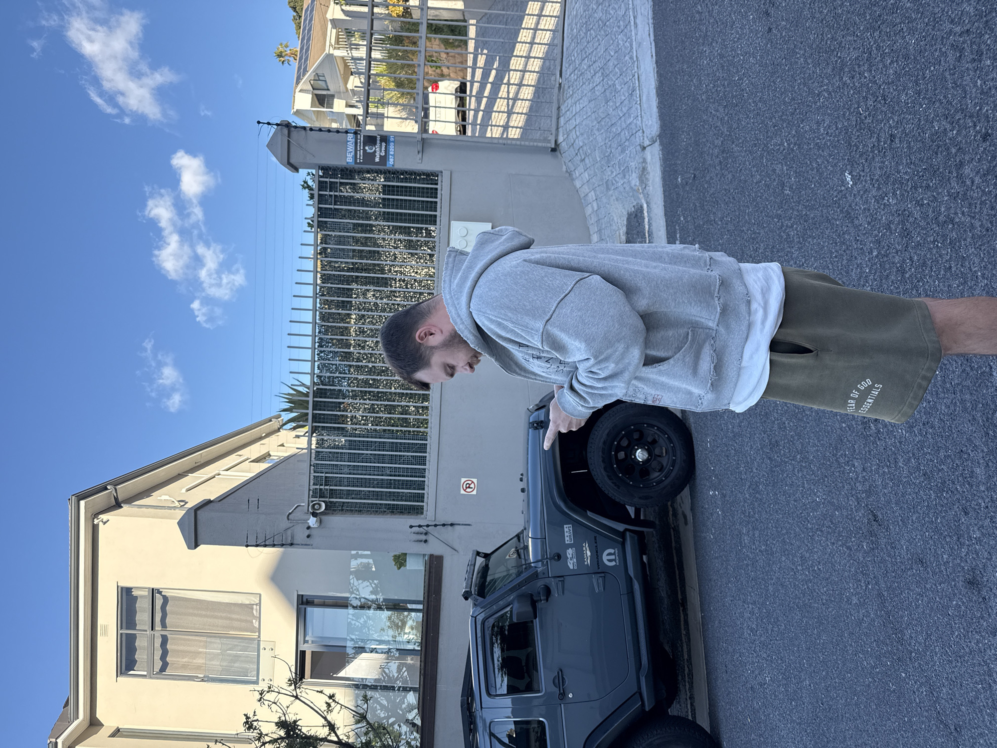 A medium shot of a person facing right. The person is standing next to a gray Jeep Wrangler Unlimited. The person is wearing a gray hoodie with a ripped hem, a white t-shirt, and green shorts. The words "FEAR OF GOD ESSENTIALS" are printed on the shorts. The person is pointing at the car’s front driver-side tire. The car has the words "WRANGLER" and "SAHARA" on it.
Behind the person and the car is a light gray building, a metal fence, and another building. A street sign is on the metal fence. In the background there is a glimpse of a secured entrance gate. The sky is visible and is blue with scattered clouds. The person is standing on a paved sidewalk and the car is parked on the road.
The location is Cape Town, South Africa.