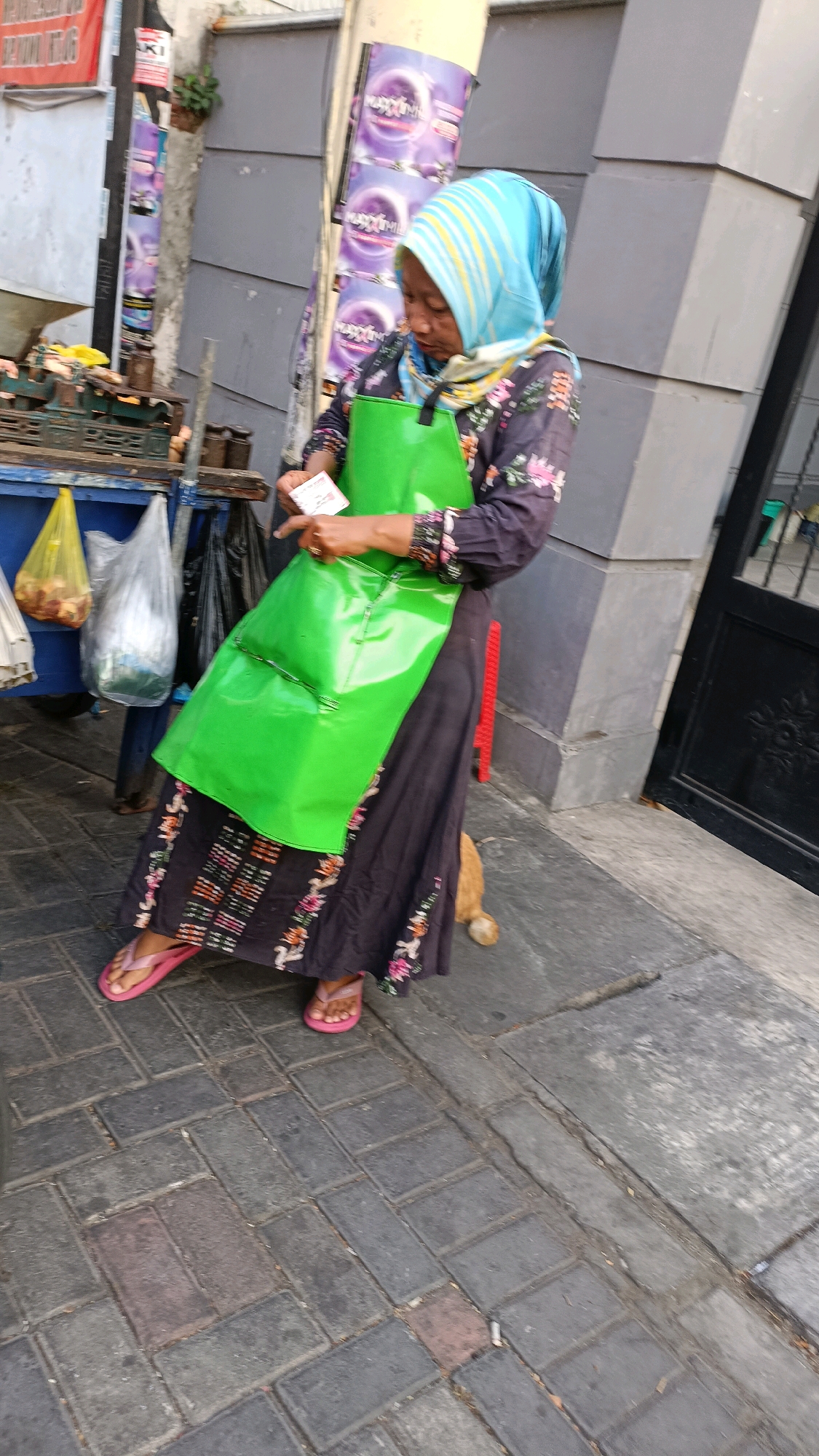 An Indonesian woman wearing a light blue and yellow hijab, a floral print blouse, and a dark skirt with floral embroidery is standing on a paved sidewalk. She is wearing pink flip-flops. She has a bright green apron tied around her waist. In her hands, she holds a small white card and appears to be showing it to someone out of frame. A tan cat is visible behind her, with its hindquarters on the pavement.
To her left, there is a blue cart with various items, including two plastic bags, one yellow and one clear, hanging from its side. The yellow bag contains what appears to be food items. Behind the woman and to her left, a grey wall with a tiled pattern is visible, along with a pole with several posters depicting purple and white imagery. The setting appears to be a street in Surabaya, Indonesia, indicated by the posters and the style of the building. The ground is paved with interlocking rectangular stones.