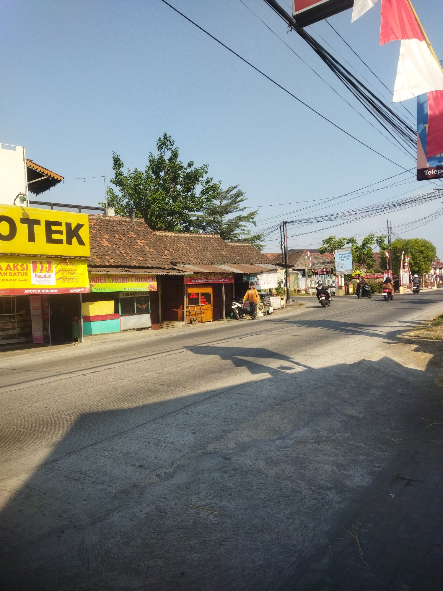 The image depicts a street scene in Juwiring, Indonesia, with a clear blue sky and buildings lining the right side of the road. A prominent yellow sign with the word "OTEK" in large black letters is visible on the left. Adjacent to it are shops with various signs, including one for "SABEN FRIED CHICKEN" and another for "NASI GORENG". Several motorcycles are present, with some being ridden by individuals on the road. One person in an orange shirt stands near a motorcycle in front of a shop. Red and white flags are displayed on poles, suggesting a national celebration or event. Power lines crisscross the upper portion of the image, casting shadows on the road. The road surface appears to be made of concrete.