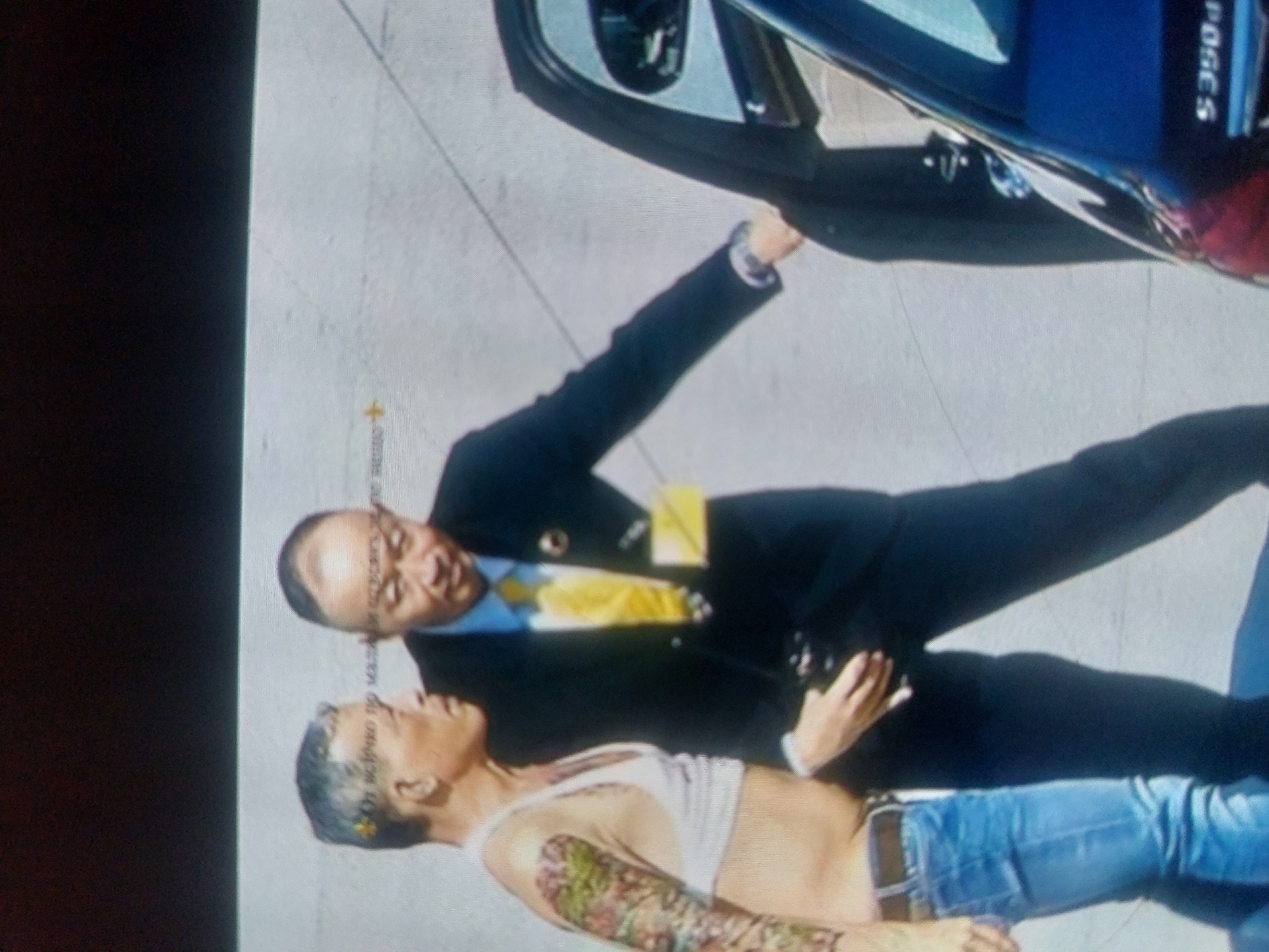 A man in a black suit and tie is standing next to a woman in a white tank top and blue jeans lying on the ground. The man is opening the door of a blue car. It is unknown whether the image was taken in Tsarevo, Bulgaria.