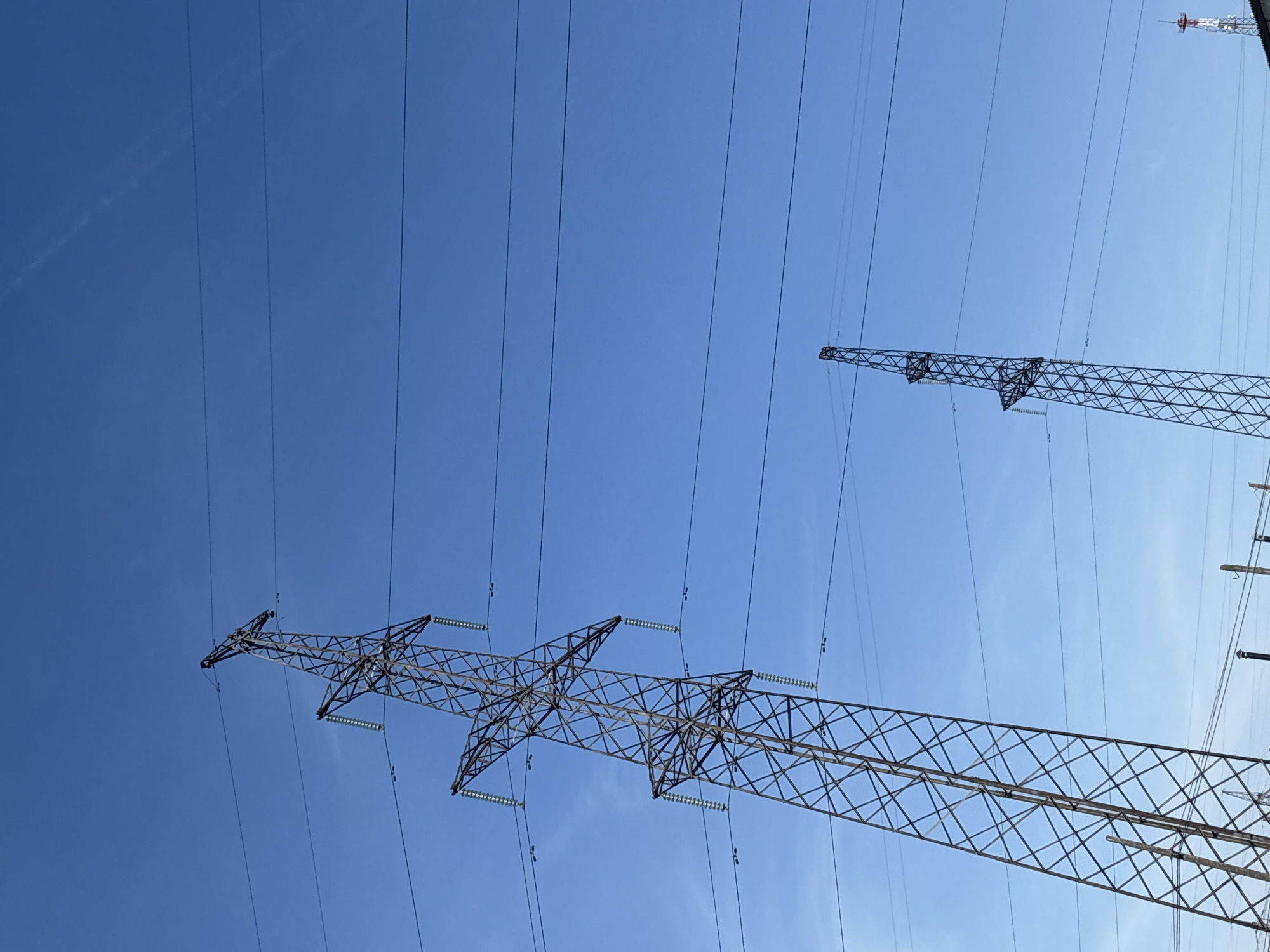 The image depicts a view looking up at high-voltage power lines and transmission towers against a clear blue sky. Two primary towers are visible, constructed of steel lattice framework. One tower is closer to the camera and extends across a significant portion of the frame. Insulators are attached to the tower to support the power lines. The other tower is further in the distance and appears smaller. Several power lines run horizontally across the sky. In the upper right corner of the frame, a cellular tower structure can be seen. This is potentially located in Chaikovsky, Russia.