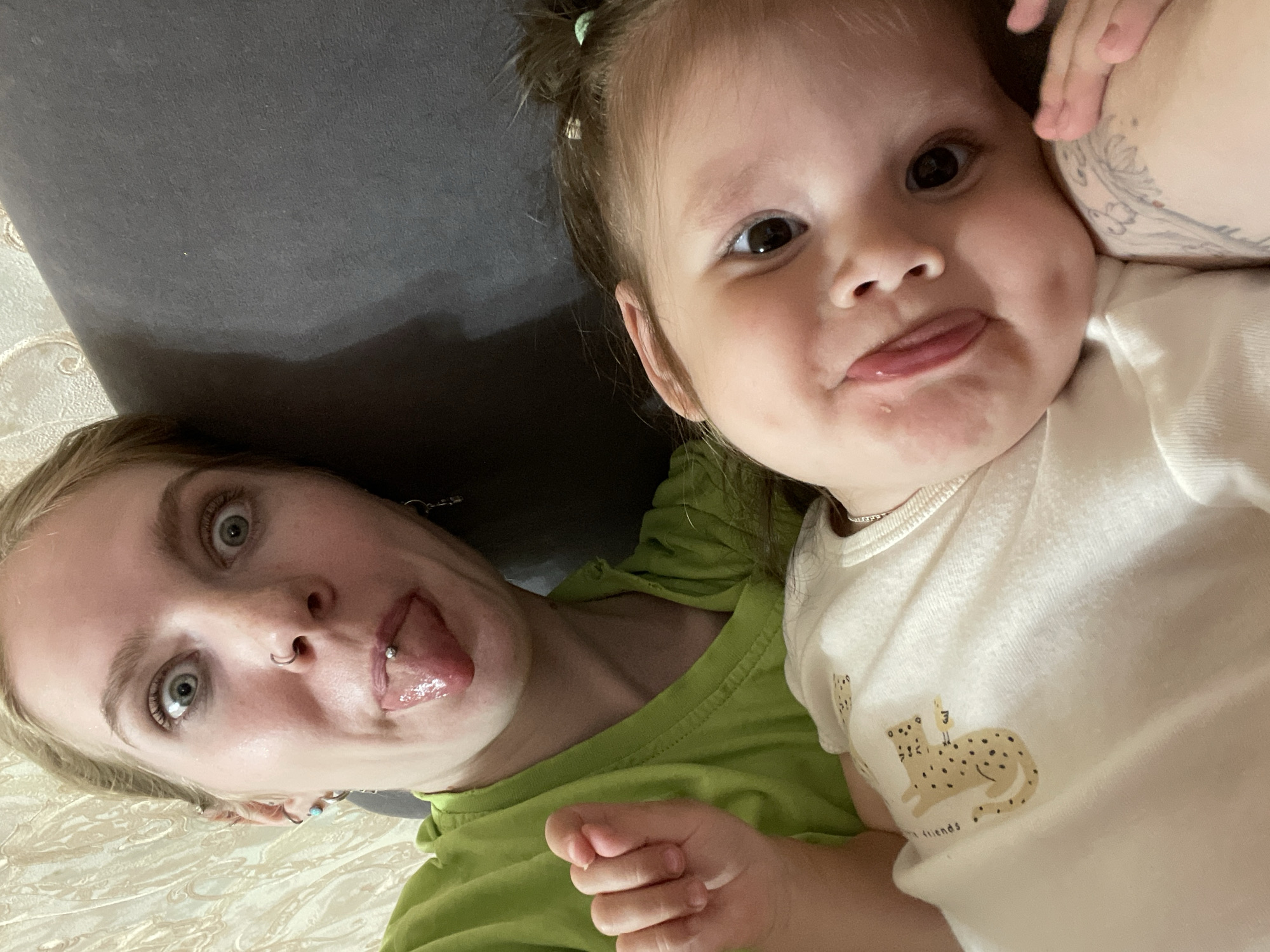 An overhead shot captures an adult woman and a young child making playful faces while lying down indoors. The woman, with light hair and distinct blue eyes, is on the left, sticking out her tongue which has a visible piercing. She also has a nose ring and wears a lime green t-shirt. Her expression is wide-eyed and humorous.
Next to her, on the right, is a toddler with dark hair tied up, dark eyes, and a round face. The child is also sticking out their tongue playfully. They are wearing a light-colored long-sleeved top featuring a printed design of a leopard or cheetah lying down, with faint text beneath it that reads "friends". A minor rash or discoloration is visible around the child's mouth and chin area.
The background consists of a dark grey, likely upholstered, surface (possibly a couch or cushion) and a light-colored, patterned fabric (like a bedsheet or blanket) that runs along the bottom left. The lighting is artificial, suggesting an indoor setting without specific time-of-day cues. The overall mood is lighthearted and affectionate. No specific external location cues are visible within the image.