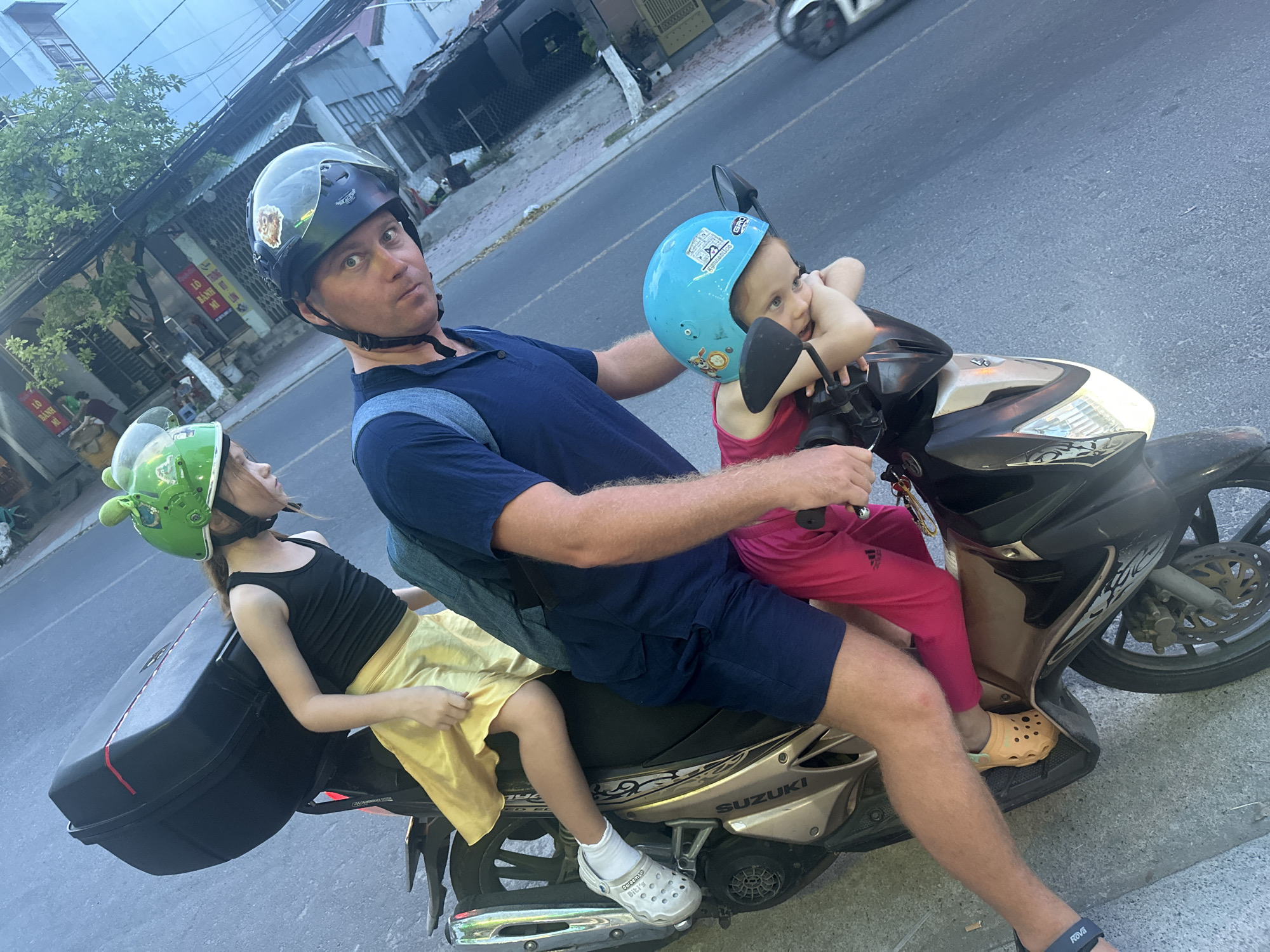 In Bac Nha Trang Ward, Vietnam, a man wearing a dark blue polo shirt, dark blue shorts, and a black helmet sits on a gold Suzuki motorcycle. He is driving the motorcycle on a road. Behind him, a young girl wearing a black tank top, yellow skirt, and green helmet sits on the back. In front of him, another young girl wearing a pink shirt, pink pants, and a blue helmet is holding onto the handlebars. The road is gray and made of asphalt. Buildings are visible on the left and right side of the road. A white vehicle is in the background. A black storage container is attached to the rear of the motorcycle. The man and children are wearing Crocs.