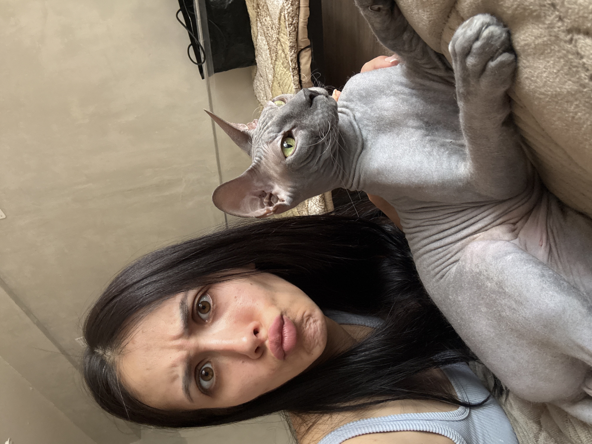 A woman with long, dark hair and brown eyes is holding a hairless cat, likely a Sphynx, close to her face. The woman is making a pouting face, with her lips pursed. The cat is gray, with green eyes, and its paws are resting on a tan-colored, textured surface, possibly a piece of furniture. The background includes a beige wall. The location is unknown, but the request mentions the city of Votkinsk, Russia.