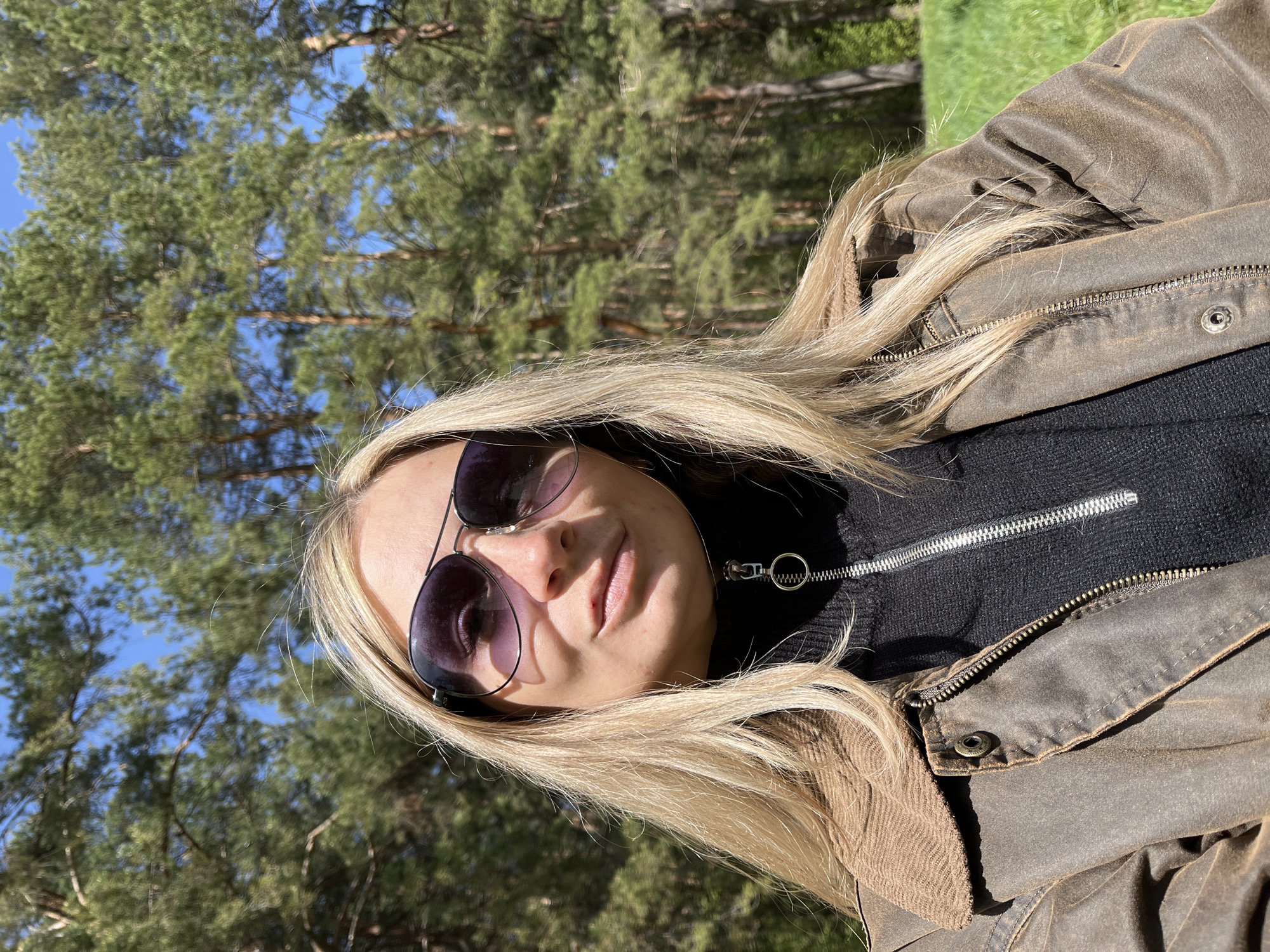 A woman wearing sunglasses, a black turtleneck, and a brown jacket stands in a forest in the city of Chaikovsky, Russia. Trees are visible in the background.