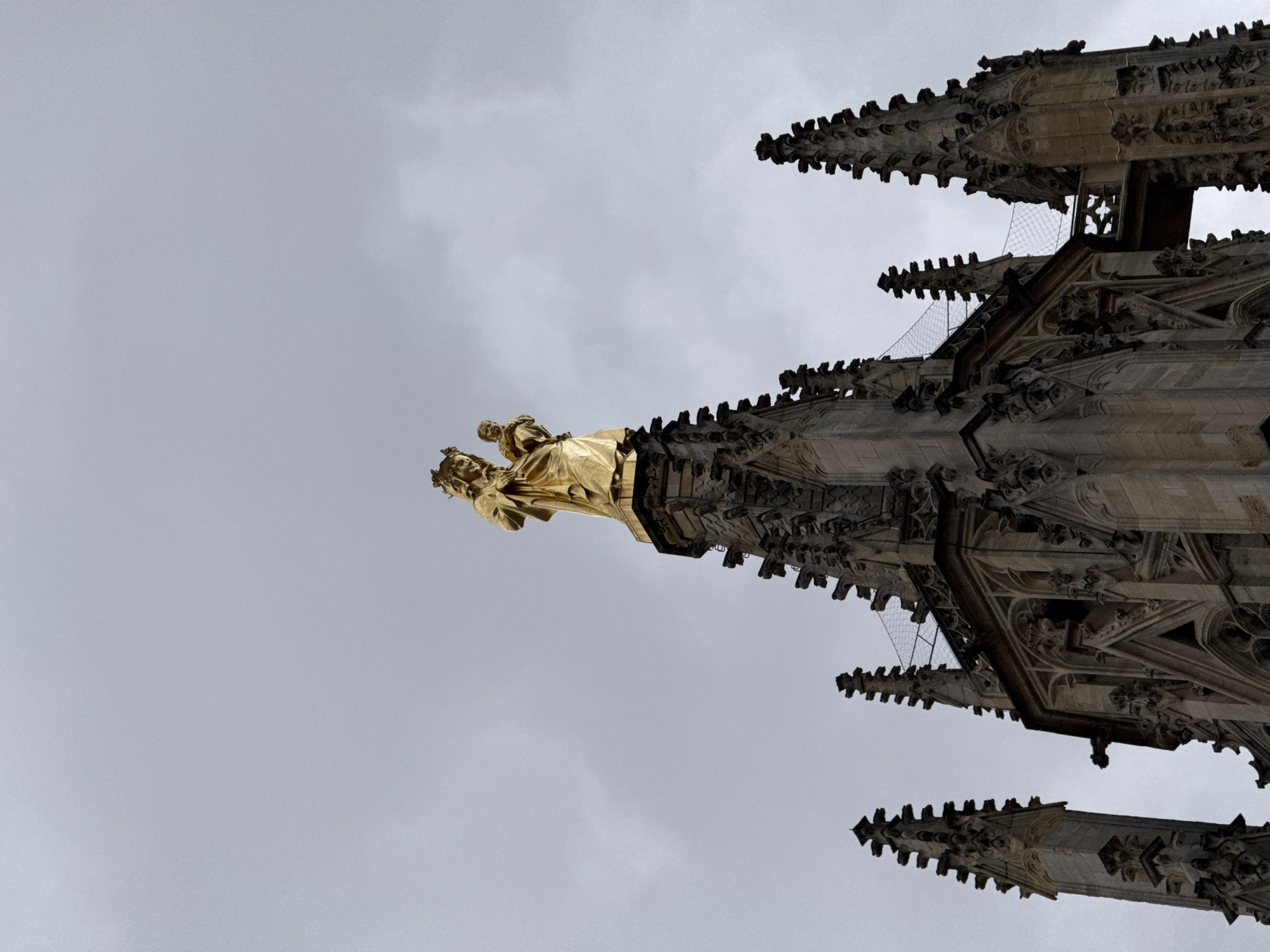 A gilded statue of the Virgin Mary holding the infant Jesus stands atop a gothic spire against a cloudy sky. The statue is positioned at the apex of a complex architectural structure, likely a cathedral or church, featuring intricate stonework, pinnacles, and gargoyles. The spire itself is surrounded by other similarly ornate gothic architectural elements, suggesting a historical religious building in a city. This specific statue, known as "La Bonne Mère," is located on the spire of the Basilica of Our Lady of Bordeaux in Bordeaux, France.