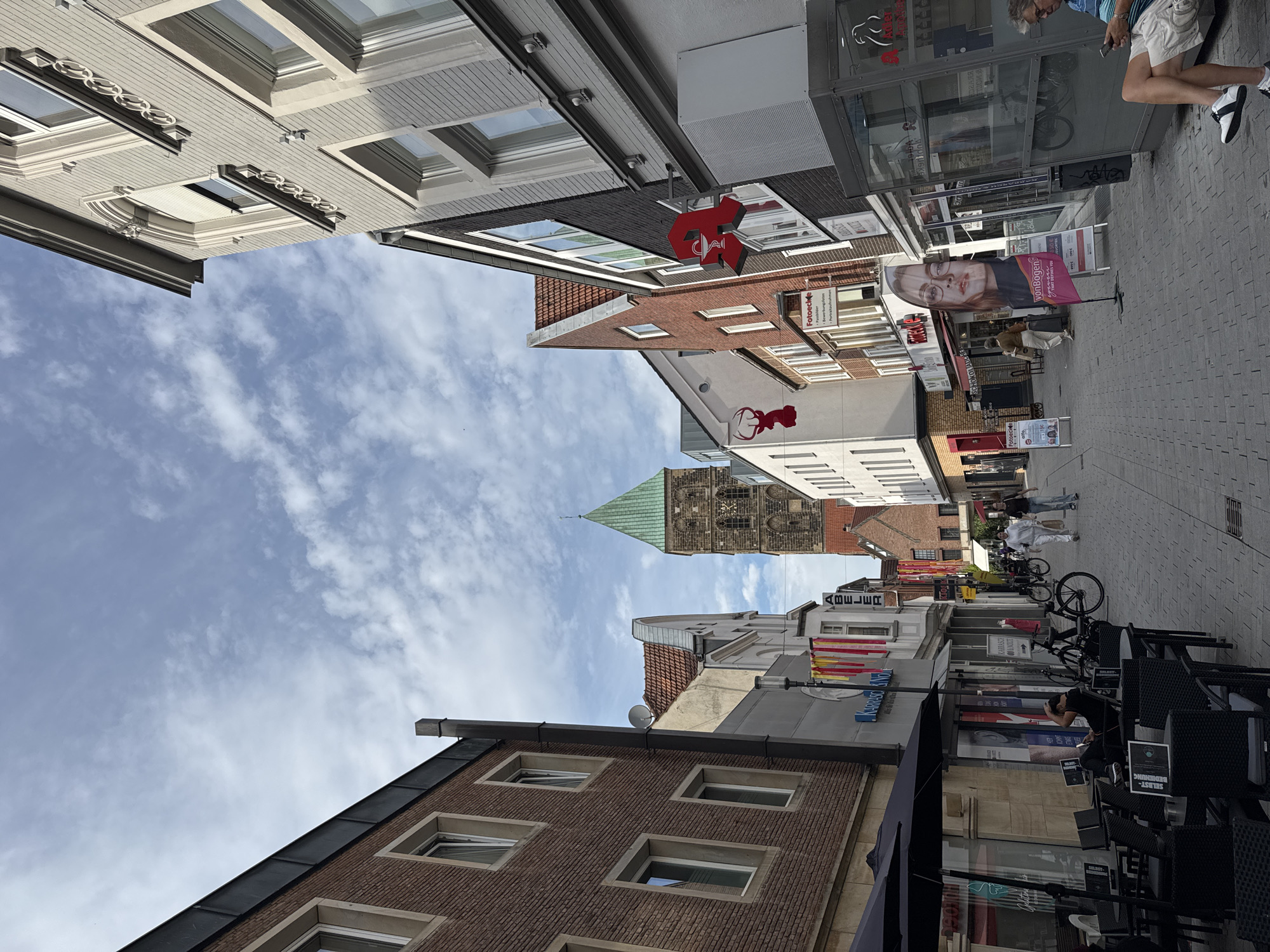 The image depicts a street scene in Rheine, Germany, likely in the city center, as indicated by the architecture and presence of shops. The perspective is from eye level, looking down a cobblestone street lined with buildings on both sides. The sky is partly cloudy, with a notable green-topped tower visible in the distance, which appears to be a church or historical structure.
On the left side of the street, there are brick buildings with multiple windows. One building has an outdoor seating area with black tables and chairs under black umbrellas, suggesting a cafe or restaurant. A man is seated at one of the tables, looking at his phone.
The right side of the street features buildings with lighter-colored facades and architectural details. Various shop signs are visible, including a prominent red pharmacy symbol, and advertising banners. One banner displays a woman's face with the text "vonBogen". Further down the street, several people are walking, and bicycles are parked.
The overall atmosphere suggests a typical European urban environment on a mild day, with commercial activity and pedestrian traffic. The presence of the specific tower and the style of the buildings are indicative of a historical German town.
