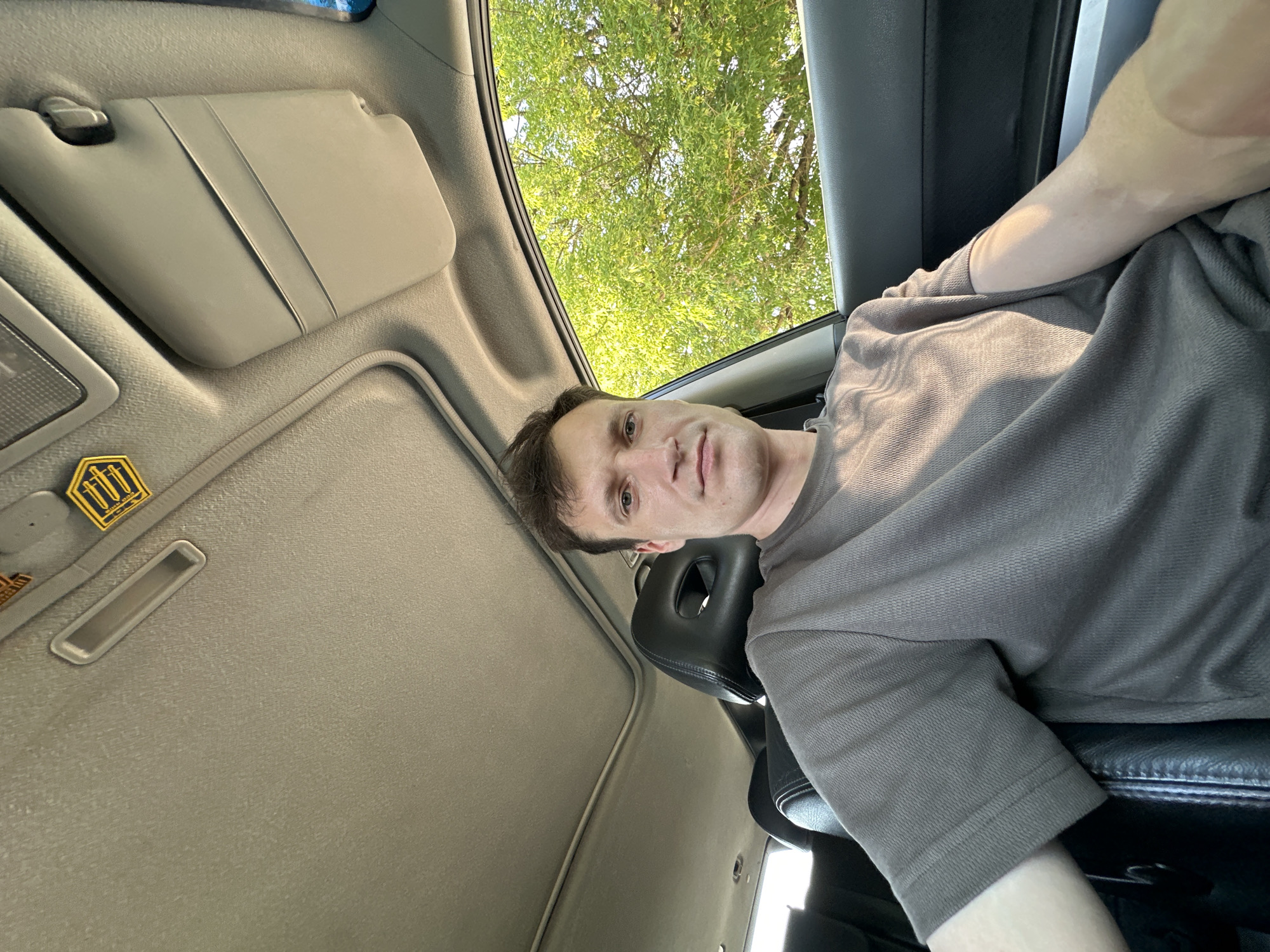 A man is seated in the driver's seat of a car. He is wearing a grey t-shirt and is looking directly at the camera. The car's interior is visible, including the sunroof and the dashboard. Outside the car, green trees can be seen. The image was taken in the city of Makiv, Ukraine.