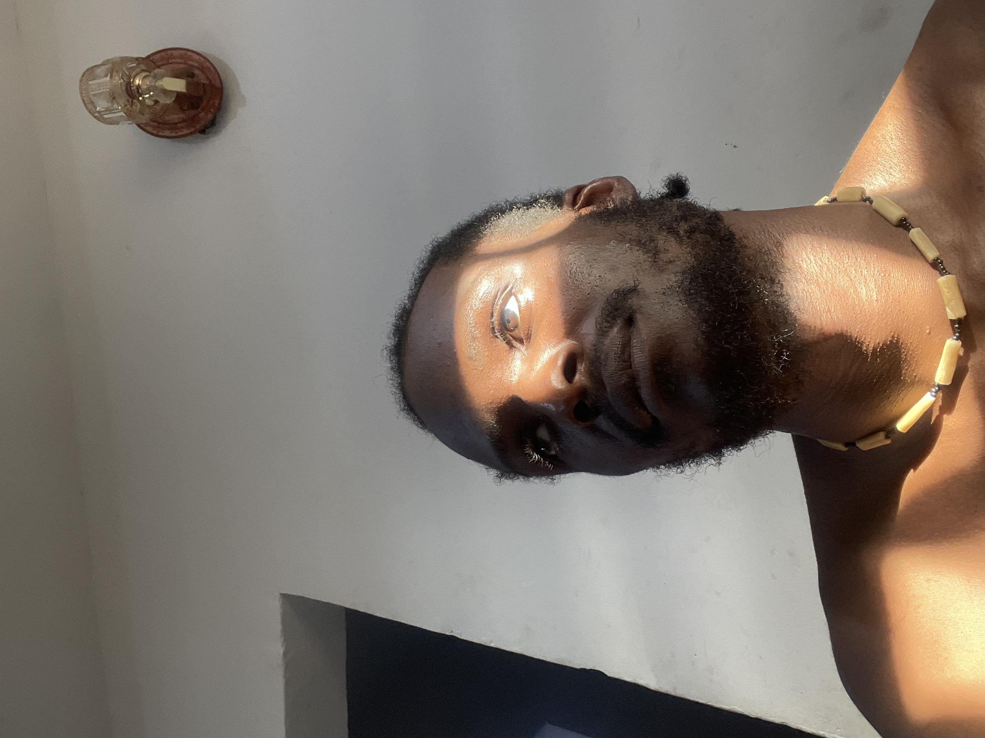 This media file is a close-up, vertically oriented photograph of a person bathed in dramatic sunlight. The subject appears to be a man with dark skin, a beard, and a beaded necklace around his neck. The light casts strong shadows, highlighting his features and creating a chiaroscuro effect. The background is a plain, light-colored wall, with a shadowed area and a hint of a dark object in the lower right corner. In the upper left corner, a small, decorative glass object, possibly a light fixture or a religious artifact, is visible. The quality of light suggests it is either early morning or late afternoon. The overall mood is intimate and contemplative, with the strong light drawing attention to the subject's face and form. There is no visible text in the image. The location context is Abuja, Nigeria.