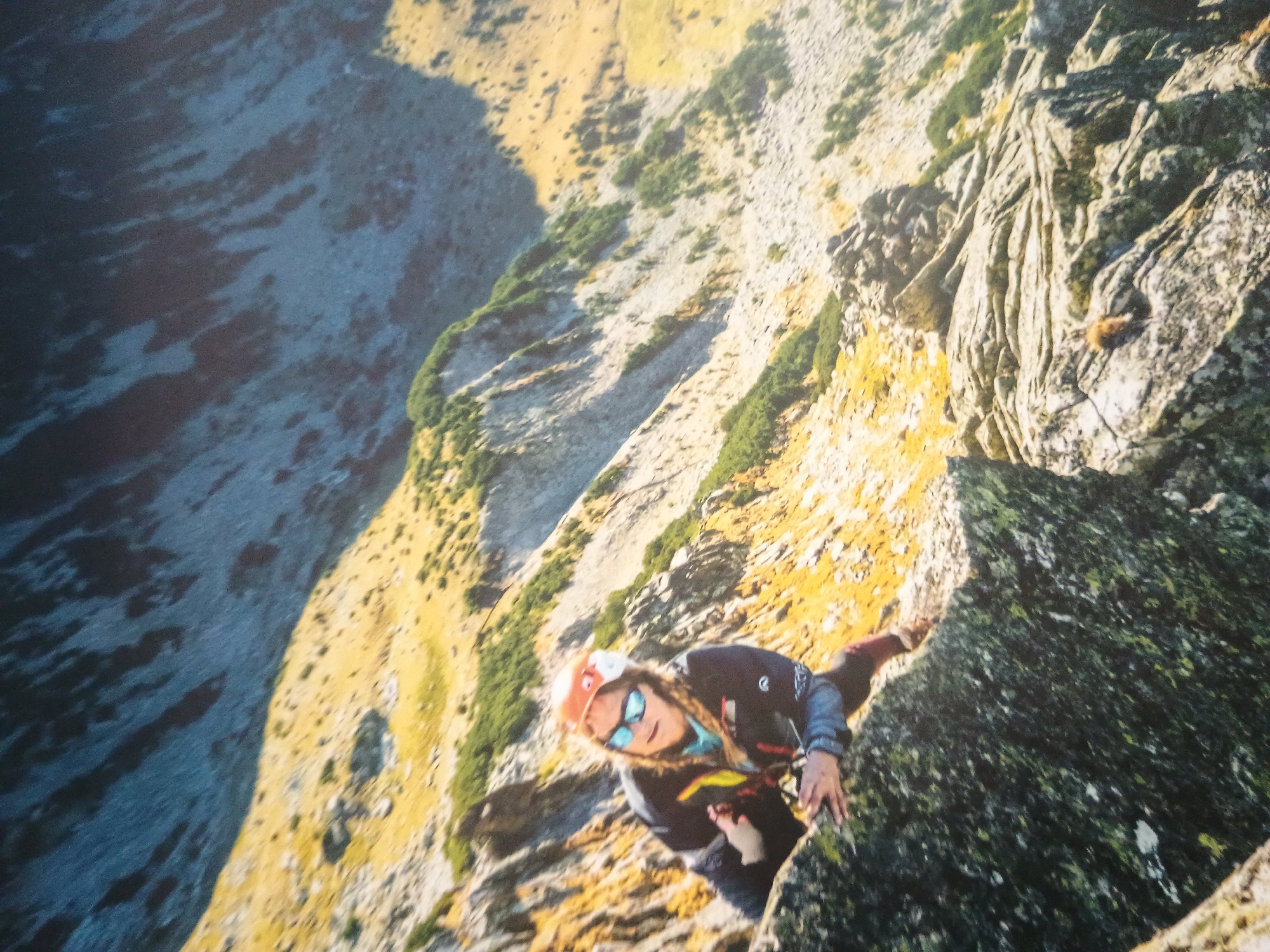 A climber in an orange helmet and sunglasses clings to a craggy rock face. The climber wears a dark jacket with red accents and gray sleeves. The rock face is varied, ranging from light gray to dark green with patches of yellow. In the background, a steep mountainside covered in sparse vegetation leads down to a gray, water-like area. The location is an unidentified mountain range within the city of Unknown, Unknown.