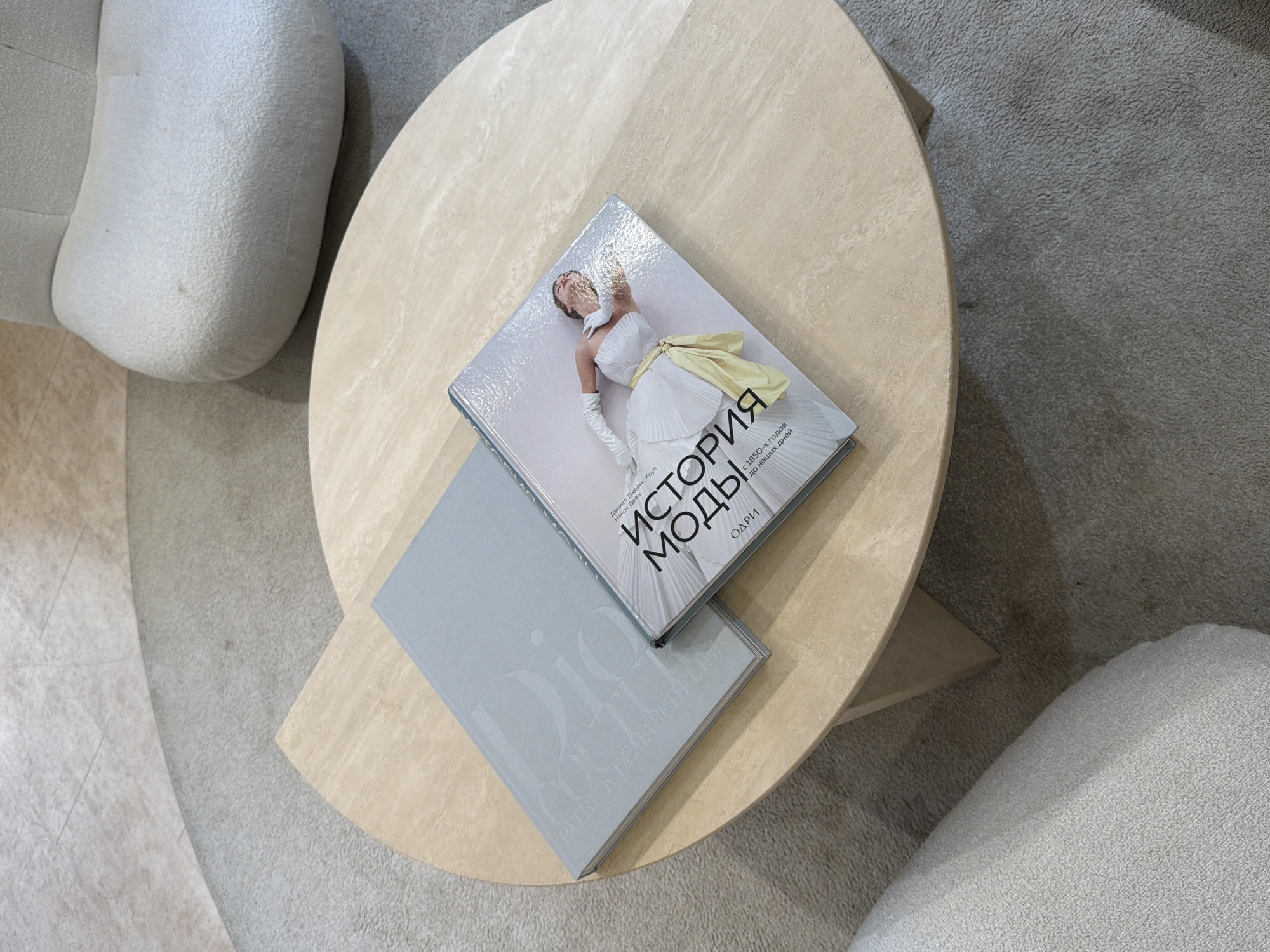 The image shows a top-down view of a modern interior setting. A beige, round coffee table with a slightly rough texture is the focal point. Two books are placed on top of the table, slightly overlapping each other. The book on top has a shiny cover with a woman in a white dress and yellow ribbon on it, accompanied by Russian text that reads "История моды" (History of Fashion). The bottom book is light grey, with the words "Dior Couture" and "Patrick Demarchelier" faintly visible. Parts of two white, soft-looking armchairs are seen on either side of the table. The floor around the table and chairs consists of beige flooring and light grey carpet. The overall scene appears to be indoors, likely in a living room or lounge area. There is no indication of a specific location within Moscow, Russia or any individuals present.