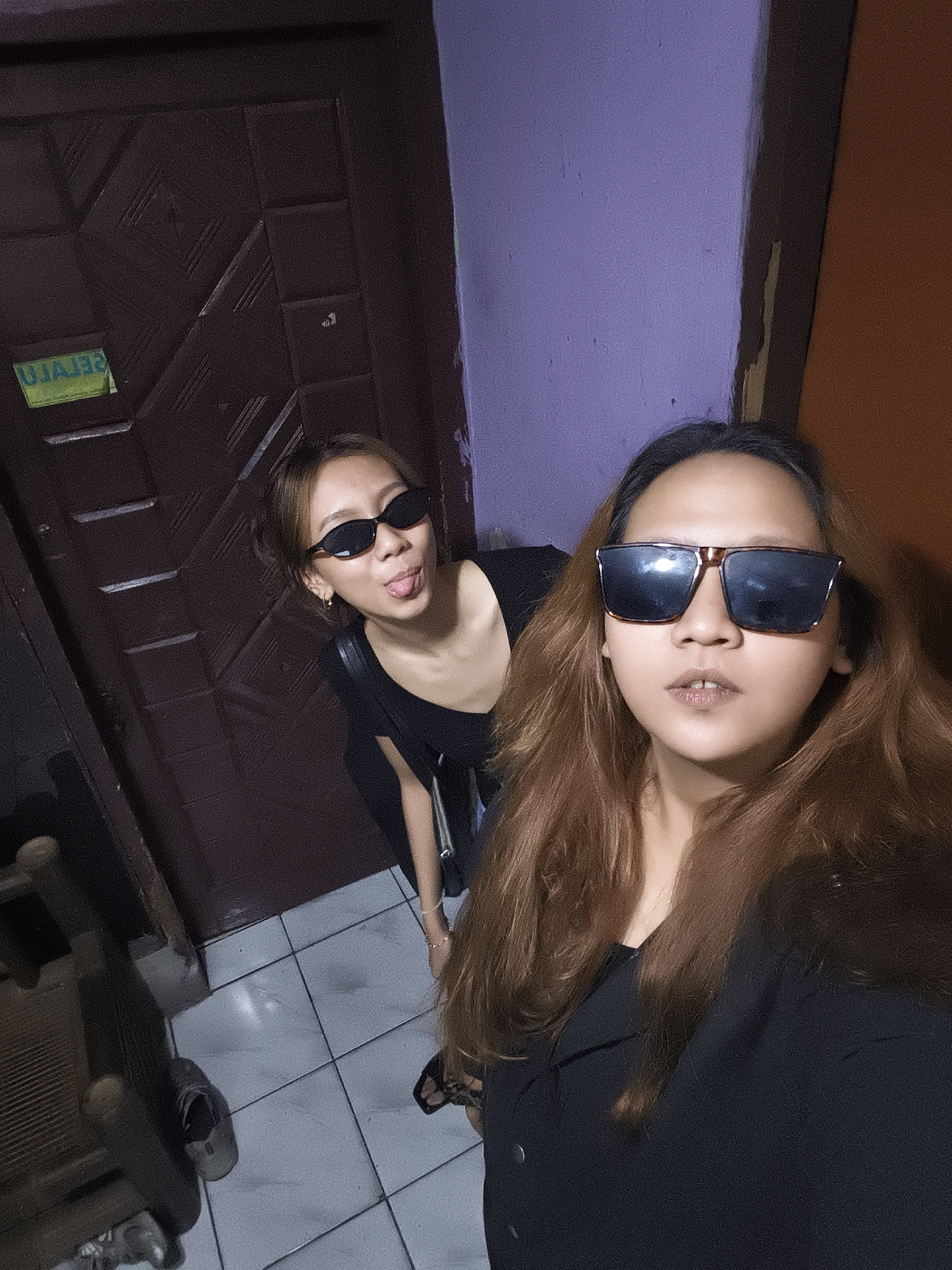 In Depok, Indonesia, two young women are captured in a selfie taken indoors. The woman on the left, positioned closer to a dark brown door with a faded green sticker, wears black sunglasses and sticks out her tongue. She has short, light brown hair and wears a black top. The woman on the right, in the foreground, wears large, square-framed sunglasses and has long, reddish-brown hair. She is dressed in a black top. The environment appears to be a hallway with tiled flooring and purple and orange walls. A portion of a wooden chair is visible in the lower left corner.