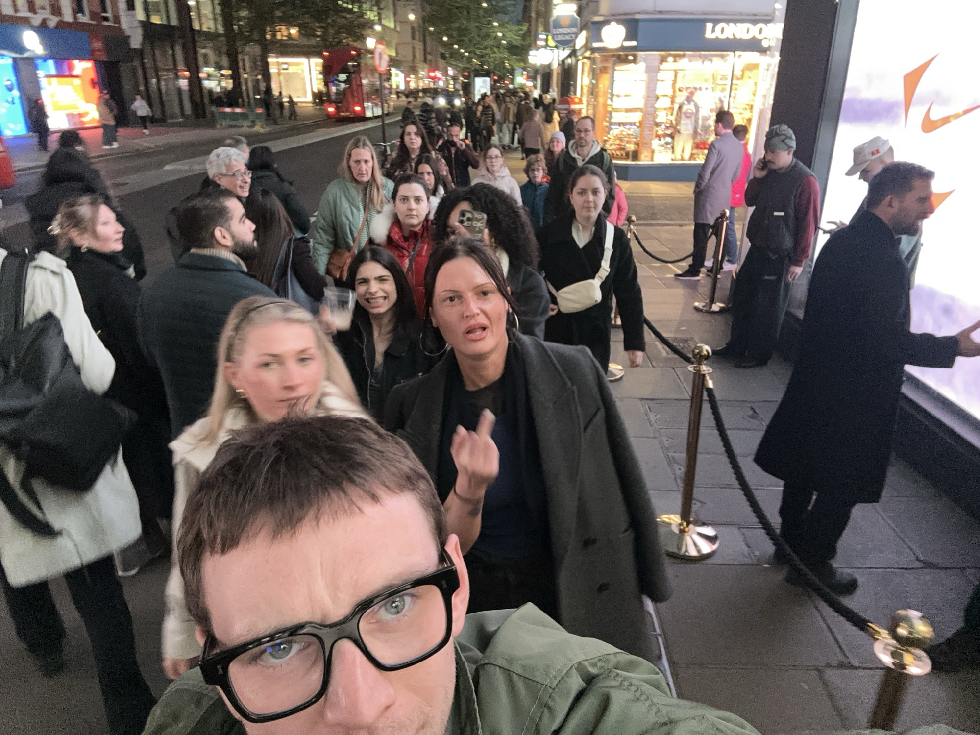 The image captures a scene in London, United Kingdom, at night. A queue of people stretches along a sidewalk, separated from the street by a rope barrier with golden stanchions. The storefront of "London Legacy" is visible behind the queue. Many individuals are casually dressed, with a mix of jackets, coats, and sweaters. The perspective suggests that the photo is a selfie, with a man wearing glasses occupying the foreground. A woman in the middle ground is gesturing with her hand. A red double-decker bus and other vehicles are faintly visible in the background, indicating a busy street. Streetlights and illuminated shop signs brighten the scene.