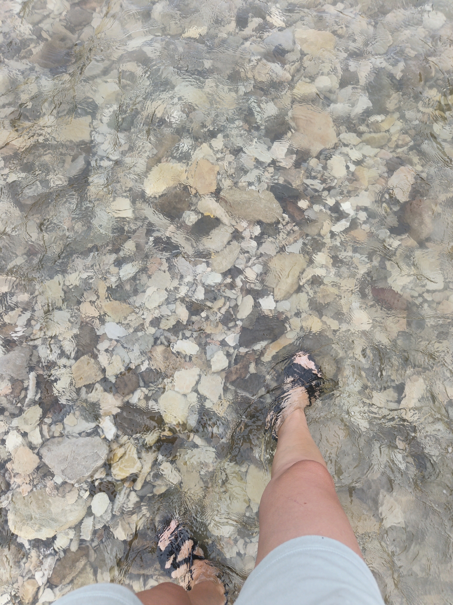 An individual's legs and feet are visible in shallow, clear water, standing on a rocky riverbed. The water distorts the view of the submerged stones, creating ripple effects. The person is wearing sandals with black and pink straps and light-colored shorts. The setting appears to be a natural outdoor environment, consistent with a river in Pshada, Russia.