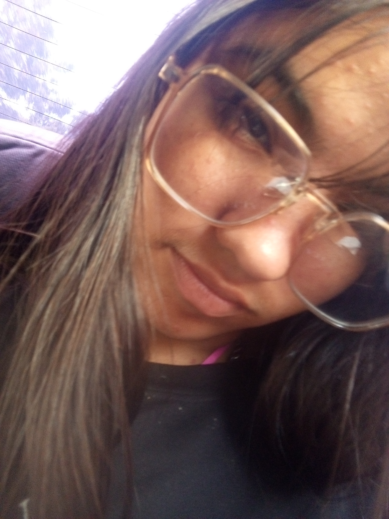 The image shows a person with long, dark hair with streaks of lighter color, wearing glasses with a light-colored frame. They are wearing a dark-colored top. The background appears to be a blurred window with horizontal lines. The location in the city of Juan Griego, Venezuela is not determinable from the image.