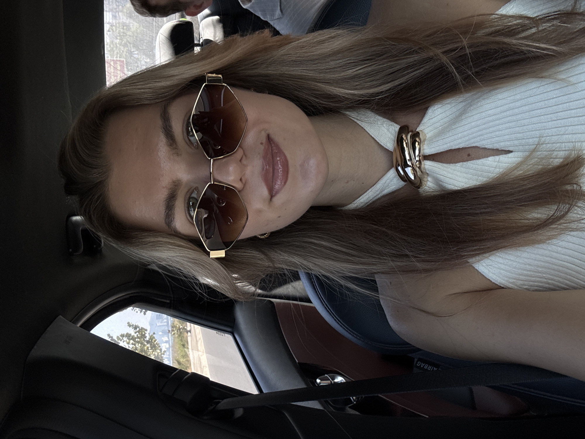 A female is photographed at close range from a low angle within a vehicle interior. The woman has long blonde hair, and is wearing octagonal sunglasses with gold frames and brown lenses. She has a light complexion, and is wearing a white ribbed top with a circular gold accent at the collar. The car interior consists of black leather seats with a brown trim. The seatbelt is visible. Outside the car window, trees can be seen. The setting appears to be an urban area, but the specific location is unidentifiable. A second individual is partially visible in the upper left corner.