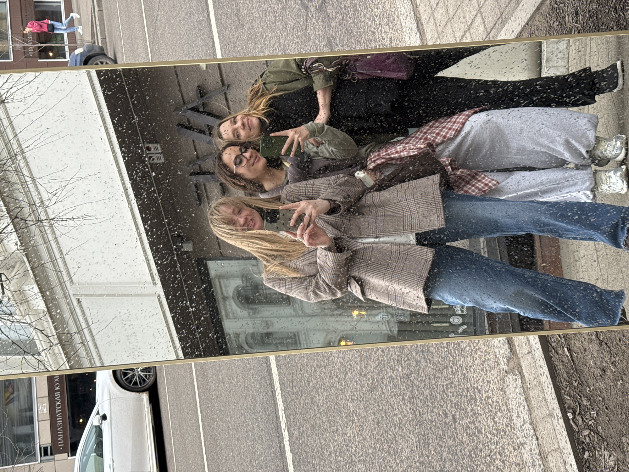 Three women are reflected in a mirror, posing and smiling. The woman at the top wears a green jacket and black pants. The woman in the middle wears glasses, a grey sweatshirt, and a red and grey plaid shirt. The woman at the bottom wears a brown plaid jacket and blue jeans. The mirror reflects a building with ornate windows. Outside the mirror's frame, parts of a white car, a street, and a building with text that includes "ПАЛАЗИА̀ТСКIЯ КУХ" are visible. In the upper left corner of the mirror, a person in a pink jacket and blue jeans is reflected. The scene is rainy. The location is Krasnoyarsk, Russia.