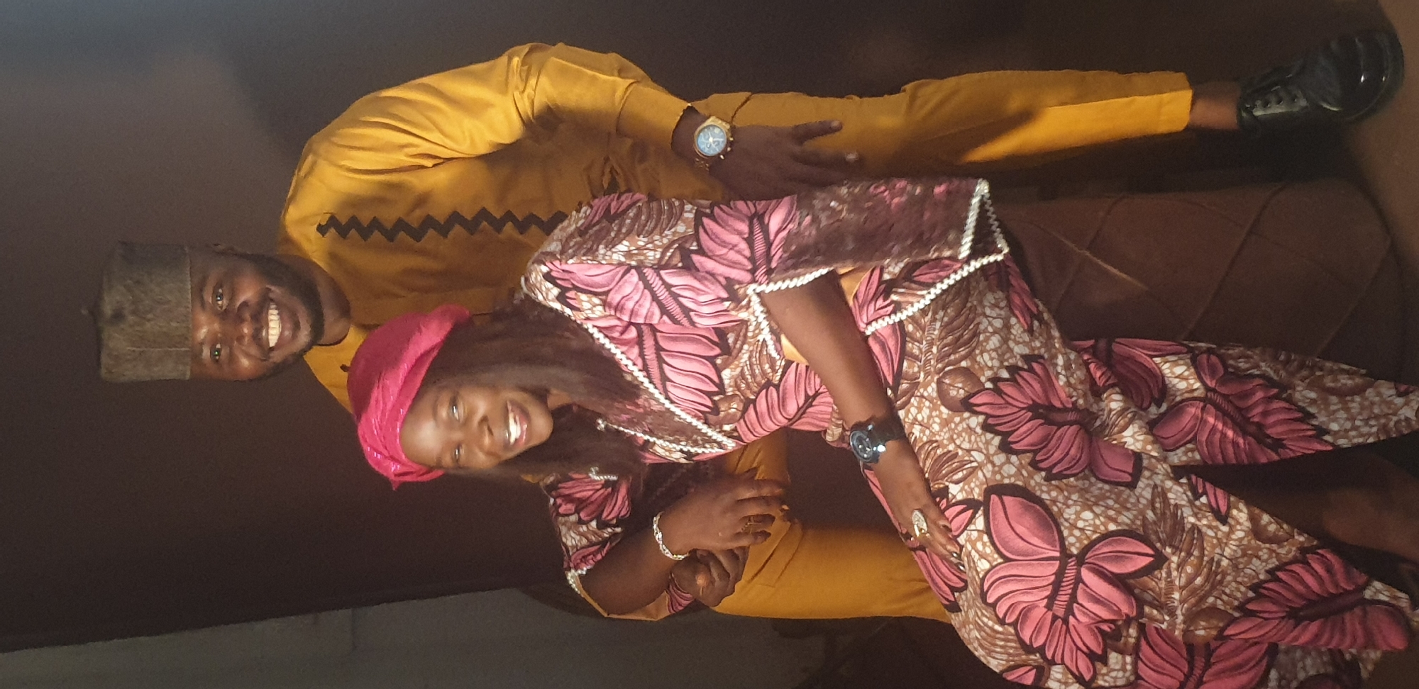 Two individuals, a man and a woman, are captured in a smiling portrait. The image is set against a dark, indistinct background, possibly indoors or in a low-light environment, with a dark brown, potentially textured surface visible behind and beneath them. The location context is Vom, Nigeria.
The man, positioned on the left, wears a mustard yellow traditional Nigerian outfit, likely a senator-style agbada, featuring a black zigzag pattern across the chest. He complements his attire with a dark, textured cap and black dress shoes. A gold-toned watch adorns his left wrist. He is smiling widely, looking directly at the camera.
The woman, standing to the man's right, wears a vibrant pink and brown patterned dress, possibly made of Ankara fabric, showcasing large floral or butterfly motifs in various shades of pink with white accents. The dress features white decorative trim along the neckline and cuffs. She has a bright pink headwrap that complements her dress and a black watch on her left wrist. A silver-toned bracelet and rings are visible on her right hand. She is also smiling broadly towards the camera, her expression conveying happiness.
The man has his right arm wrapped around the woman's waist or lower back, indicating a close or affectionate pose. Both subjects exhibit joyful emotions. There is no visible text in the image.