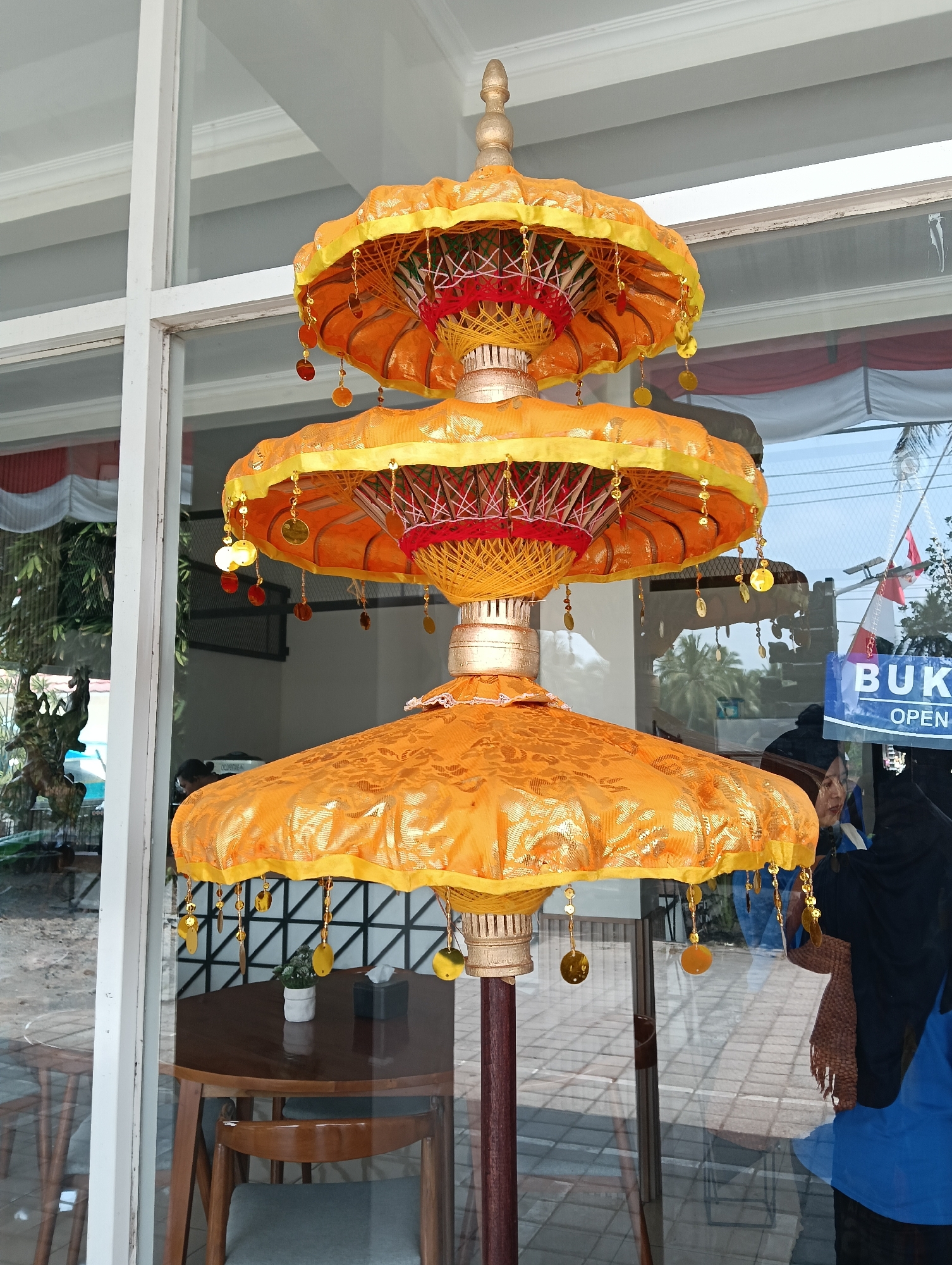 A three-tiered, golden-yellow tiered umbrella decoration is displayed vertically, in front of a window. The umbrella has intricate patterns and is adorned with small gold, red, and orange hanging ornaments. The scene appears to be an establishment in Ngrajek, Indonesia, as indicated by a "BUKA OPEN" sign visible in the reflection on the window. Inside the establishment, several people are visible, some of whom are wearing headscarves. The background outside the window shows greenery, a table with chairs, and tiled flooring.
