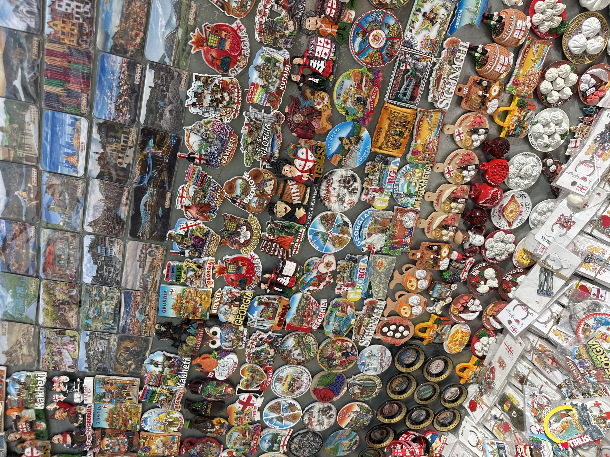 A high-angle, close-up shot shows a collection of tourist souvenir magnets, possibly at a market or shop in Tbilisi, Georgia. The magnets are predominantly of locations in Georgia with a variety of shapes, sizes, and designs. Some show images of architecture, landscapes, cityscapes, and cultural items. Several magnets feature the word "Georgia" and names of Georgian cities or regions. Many include elements of Georgian cuisine, specifically dumplings or pomegranates. Others contain cultural symbols. The arrangement is dense, with items overlapping, indicating a wide selection for potential buyers.