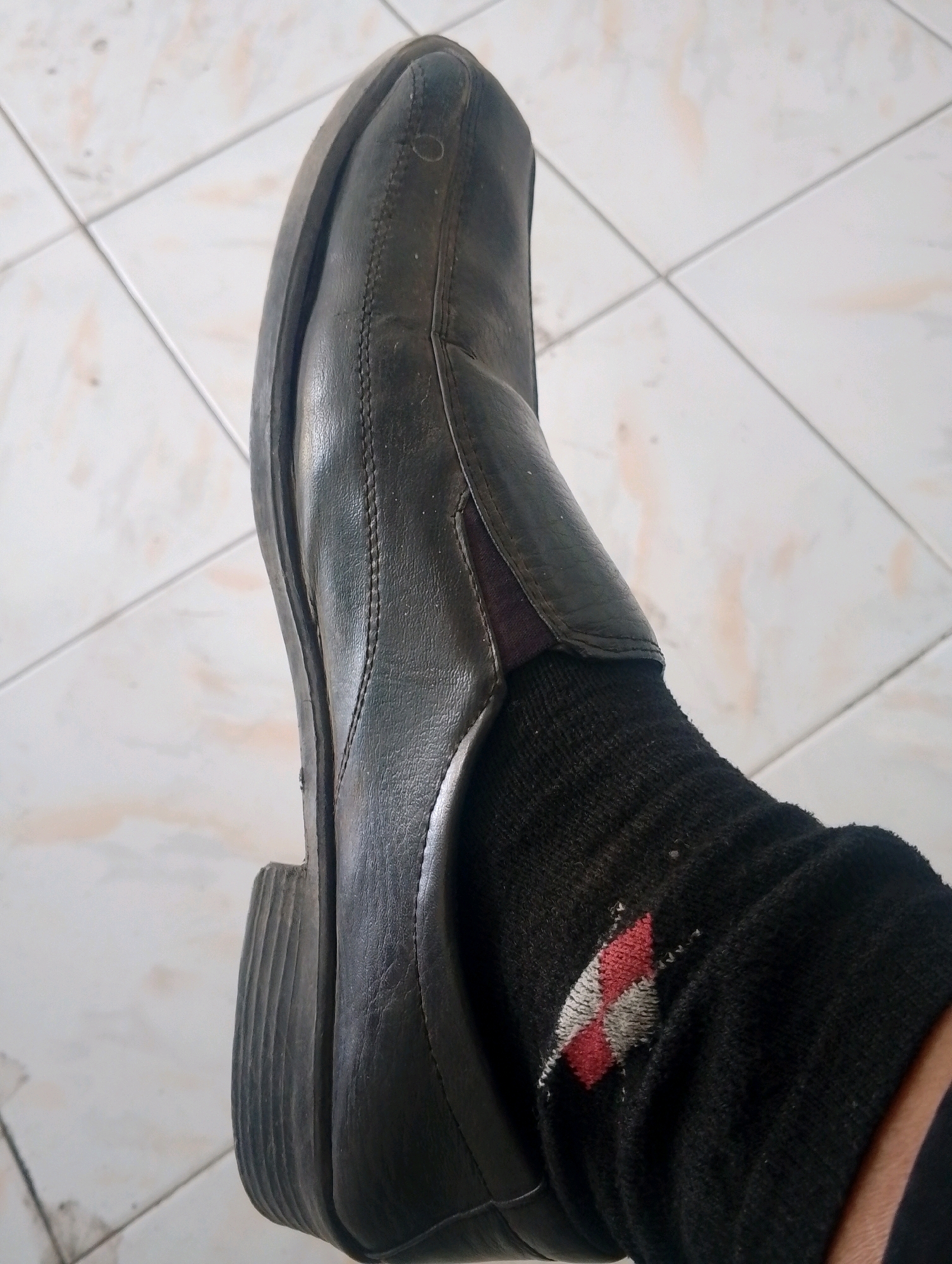 A close-up, high-angle shot shows a person's left foot wearing a black leather loafer and a black sock with a diamond pattern of red and white. The shoe appears worn, with a slight gap between the sole and the upper. The sock is pulled up the ankle and exhibits some wear and tear. The background is a tiled floor with a light-colored, diamond pattern. The scene is indoors, likely in a residential setting, given the tiling. The location is identified as Papungan, Indonesia.