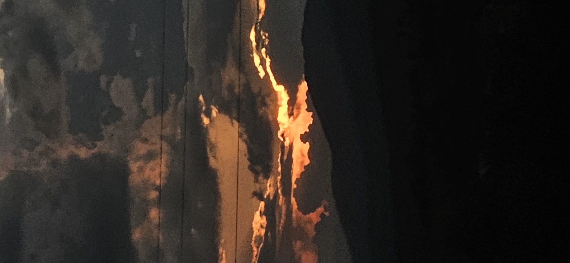 This abstract image presents a striking contrast between dark, shadowed areas and intensely bright, fiery hues of orange and yellow. The composition is vertically oriented, featuring irregular, organic shapes of light that appear to be shining through or reflecting off a textured, dark surface.
On the left and central parts of the image, the dark areas show subtle variations in tone, resembling clouds or textured material, with some faint vertical lines running through them. These dark sections serve as a backdrop for the brilliant, luminous streaks and patches of orange and yellow, which dominate the central axis and extend towards the right. The light has an almost molten, glowing quality, suggesting a powerful source such as sunlight breaking through an obstruction, reflections, or perhaps a distant fire. The rightmost section of the image is almost entirely black, indicating a deep shadow or an unlit void.
There are no discernible people, conventional objects, or explicit activities taking place, rendering the scene largely interpretative. The intense light and deep shadows evoke the dramatic lighting of either a sunrise or sunset. The image conveys a mood of mystery, warmth, and raw intensity through its vibrant color contrasts. No text is visible. The location context provided for this media file is Momin Prohod, Bulgaria.