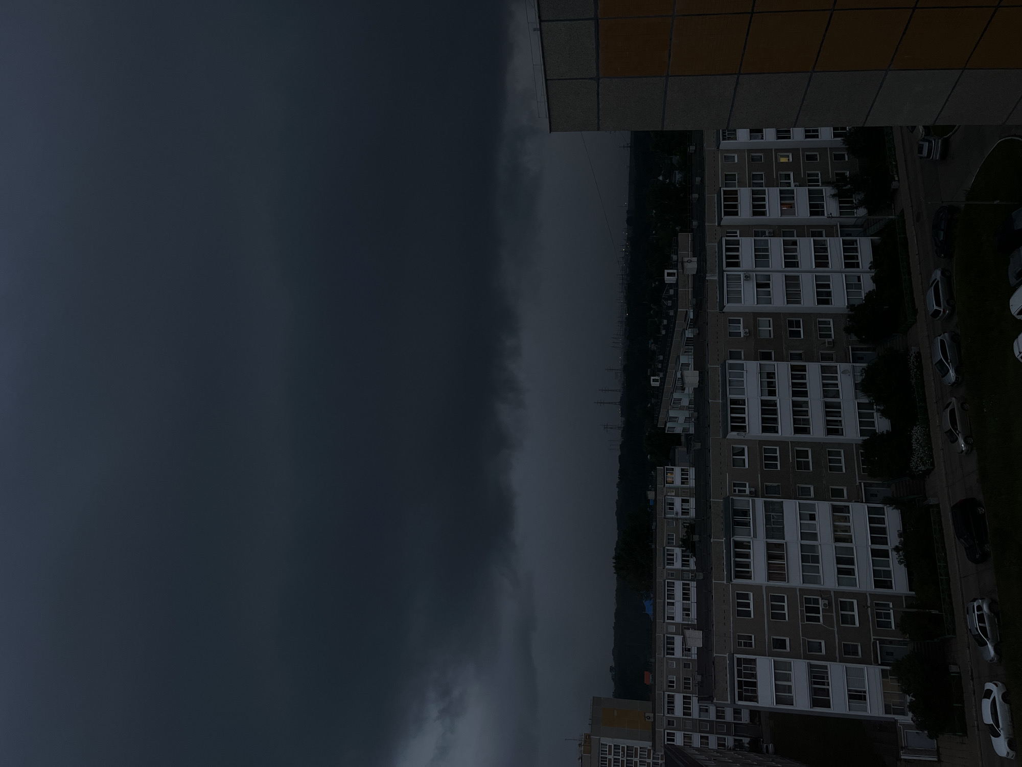 This image captures a dramatic scene from Chaikovsky, Russia, likely taken from a high vantage point, possibly a balcony or window of an apartment building. The primary subject is the stark contrast between a heavily clouded, dark sky and the urban landscape below.
The sky dominates the left side of the frame, appearing foreboding with dense, dark grey clouds that suggest an approaching storm or heavy rainfall. The line between the dark sky and the horizon is sharp, with the right side of the image showing a dimly lit cityscape.
The right side of the image displays a long, multi-story apartment building with a repetitive facade of windows. Some windows are illuminated from within, casting a warm glow that contrasts with the bleak exterior conditions. The building is situated alongside a road where several cars are parked, adding to the sense of a residential area. Trees and greenery are interspersed, suggesting a park or a planted area. The overall setting is that of a typical residential neighborhood.
The time of day appears to be late afternoon or early evening, given the low light and the dim illumination of the sky. The weather is clearly overcast and possibly stormy, contributing to a somber or dramatic mood. There are no visible people, but the presence of cars and lit windows indicates human habitation.
Notable details include the texture of the building's facade and the tiling on the top edge of the frame, which might be part of the building from which the photo was taken. The sharp demarcation of the clouds suggests a significant weather front moving through. No visible text is present in the image.