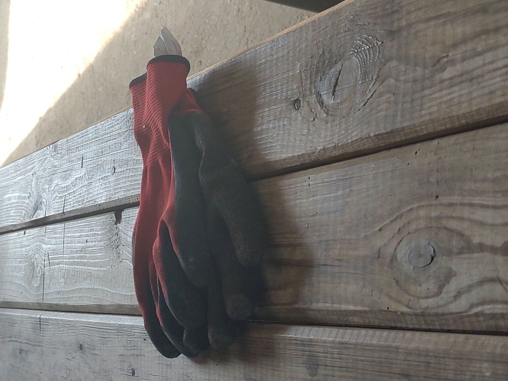 A pair of red and black work gloves is resting on a weathered wooden picnic table in Kochmar, Bulgaria. The gloves are folded on top of each other, with the red fabric of the palms and fingers visible. The black fabric of the back of the hands and cuffs is also apparent. The wooden table has a rough texture, with visible grain and some wear and tear. The background is blurred, but appears to be an outdoor setting, possibly a park or a yard, with a sandy or gravelly ground and some sunlight casting shadows.