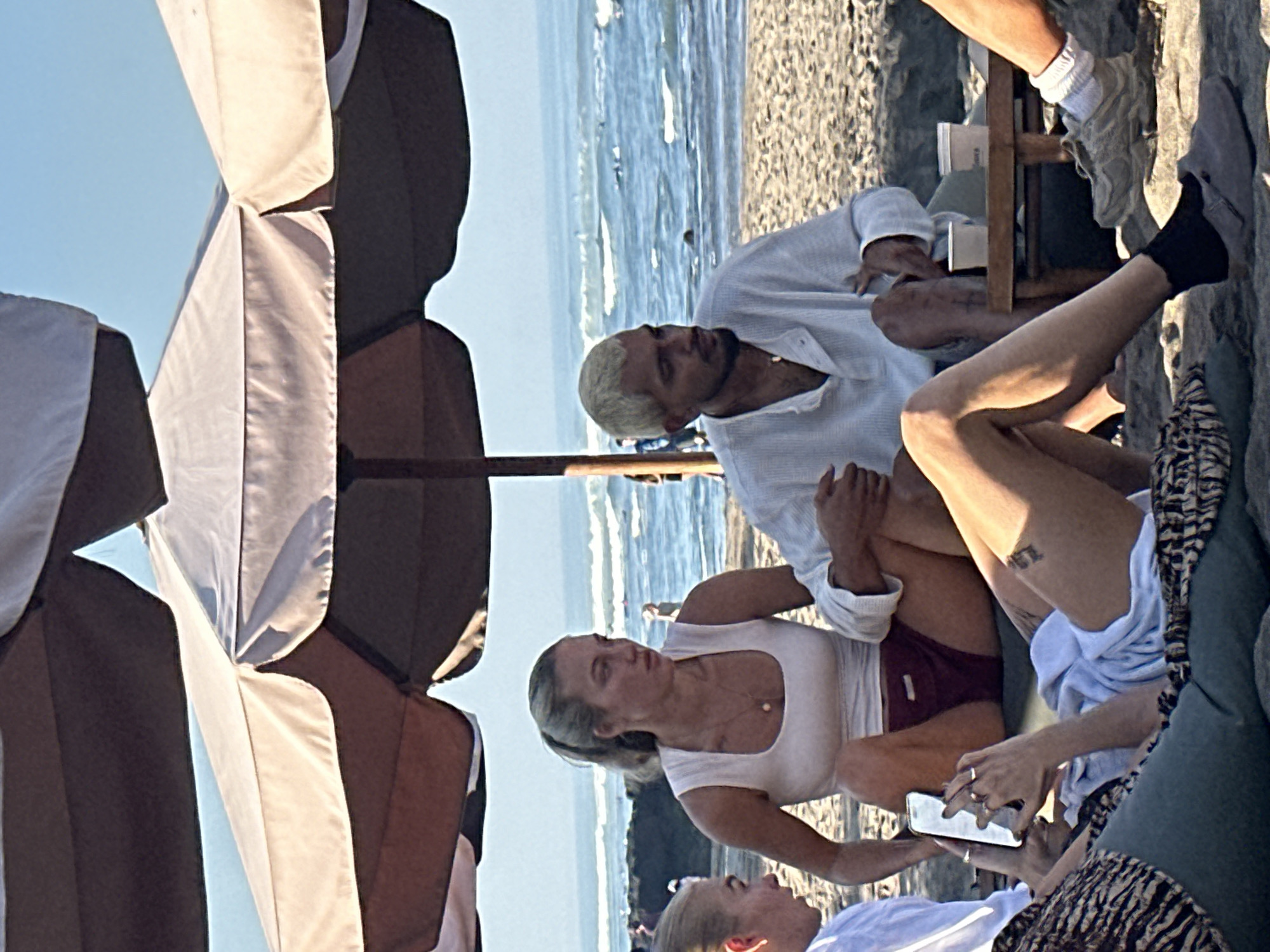 The image shows a group of people relaxing on a beach under a large umbrella. The location is specified as North Kuta, Indonesia. In the foreground, a woman is seated on the sand, holding a smartphone and looking at it. She is wearing a white top and burgundy shorts, and a tattoo is visible on her thigh. To her left, another person is partially visible, also looking towards the phone. Behind them, a man with bleached blonde hair is seated, wearing a white long-sleeved shirt. He is looking to his right. A woman with her hair in a ponytail is seated between the man and the woman with the phone, looking to her left. The background shows the ocean with waves and a sandy beach. Several other people are visible in the distance. There are two white cups on a small wooden table in front of the man. The overall scene depicts a casual beach outing.