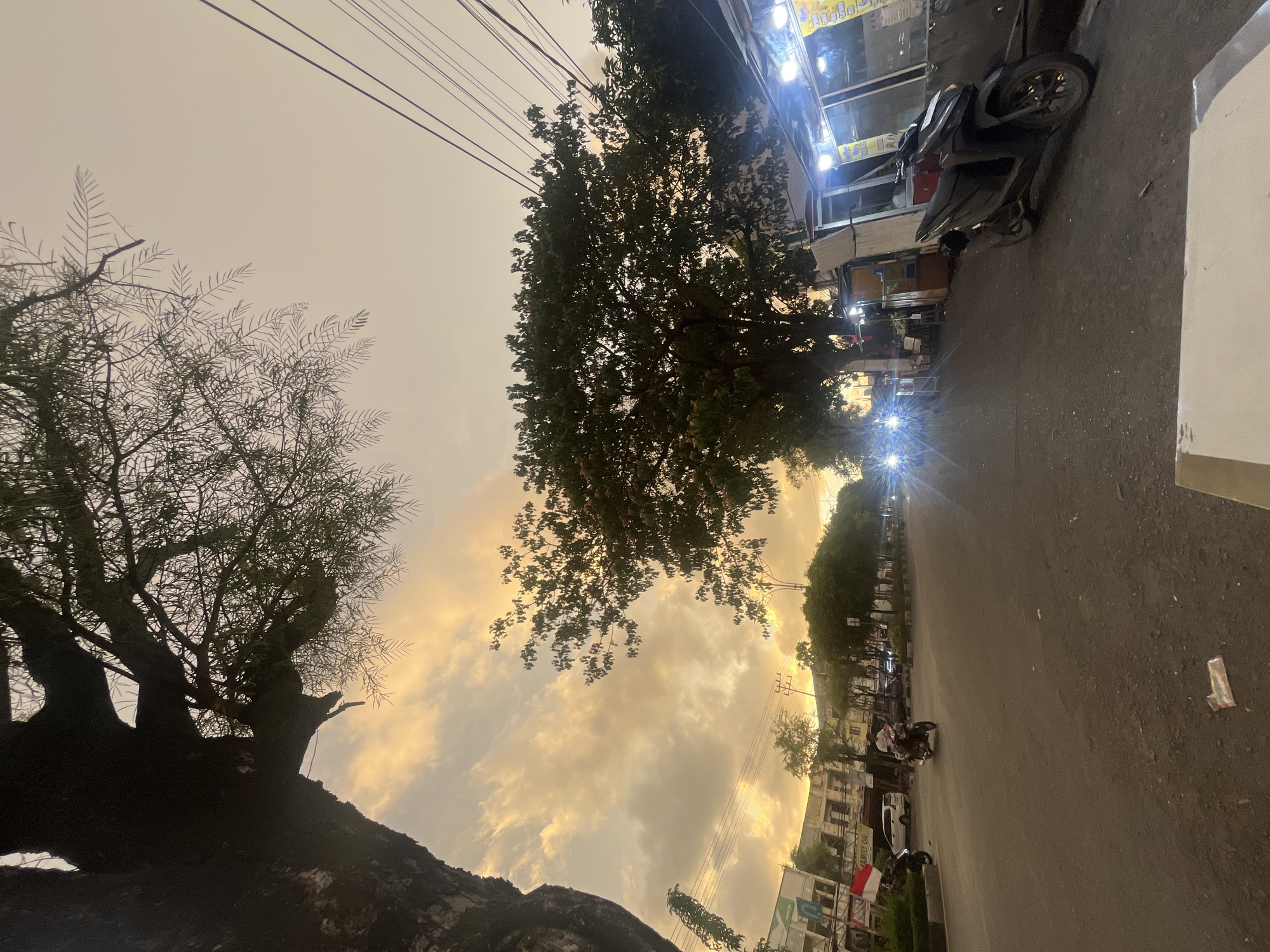 The image depicts a street scene in Banda Aceh, Indonesia, during what appears to be either sunrise or sunset. A large tree trunk dominates the left side of the frame, its branches extending upwards and to the right. The sky is filled with warm, diffused light and scattered clouds, suggesting a transitionary period of the day.
The street itself is paved and appears to be a moderately busy thoroughfare. In the mid-ground, several vehicles are visible, including a motorcycle with a rider, a white car, and other cars with headlights on, suggesting that visibility is reduced due to the ambient light conditions. On the right side of the street, there are several small storefronts or stalls with awnings and bright artificial lighting, some of which appear to be food vendors. A dark-colored motorcycle is parked in front of one of these stalls.
On the left side of the street, there are buildings, some with signage, and trees lining the sidewalk. A flag, appearing to be the Indonesian flag, is visible mounted on a pole in front of one of the buildings. The overall atmosphere is that of an urban environment at dusk or dawn, with a mix of natural light from the sky and artificial illumination from the shops and vehicles. The ground in the foreground appears to be a surface with some debris scattered on it.