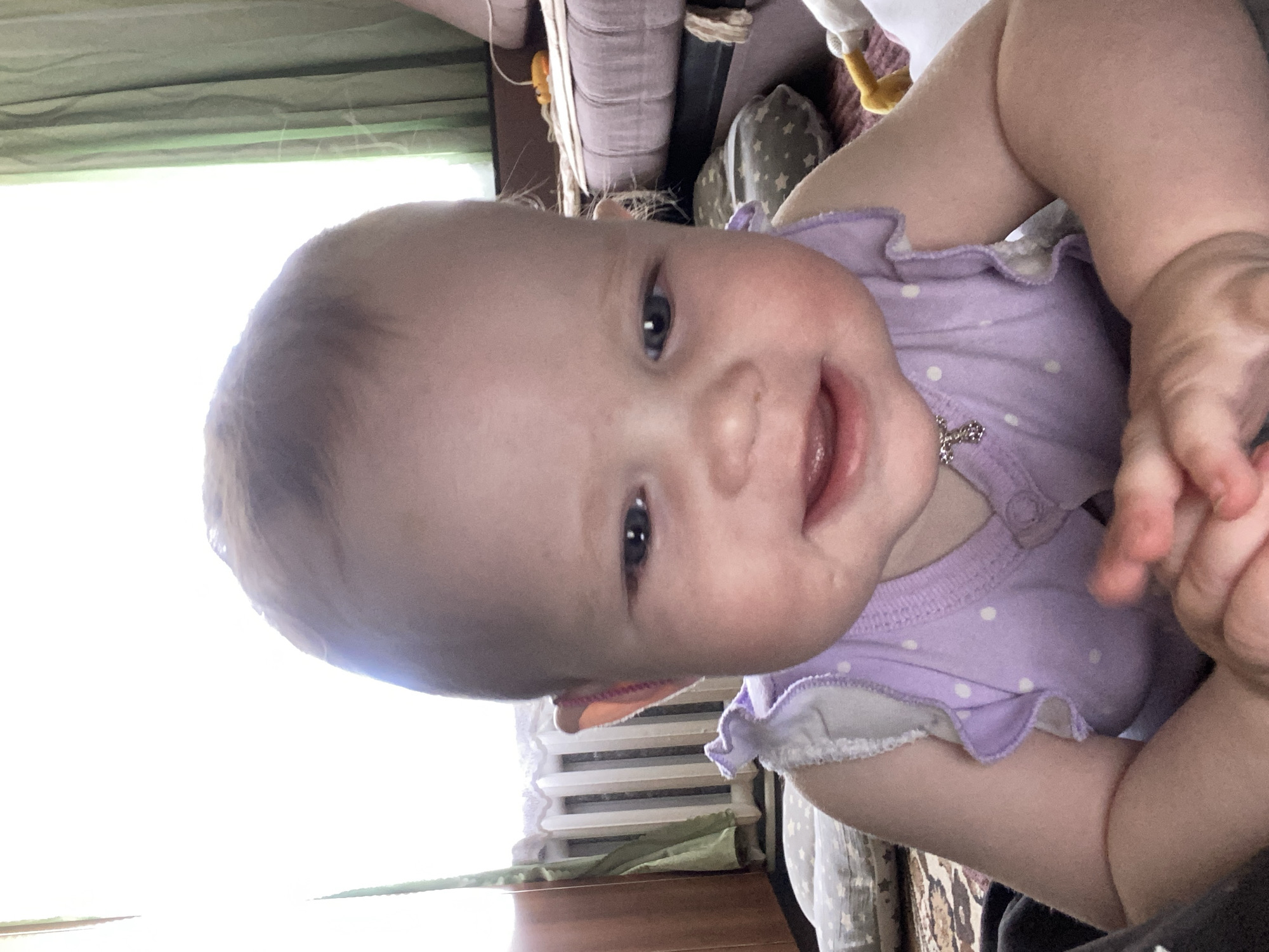 A baby wearing a light purple polka-dotted ruffled shirt is looking at the camera and smiling. The baby's skin is fair, and they have light-colored hair. The background is somewhat cluttered, featuring a window, a radiator, and a cushioned surface. The location is not discernible from the image.