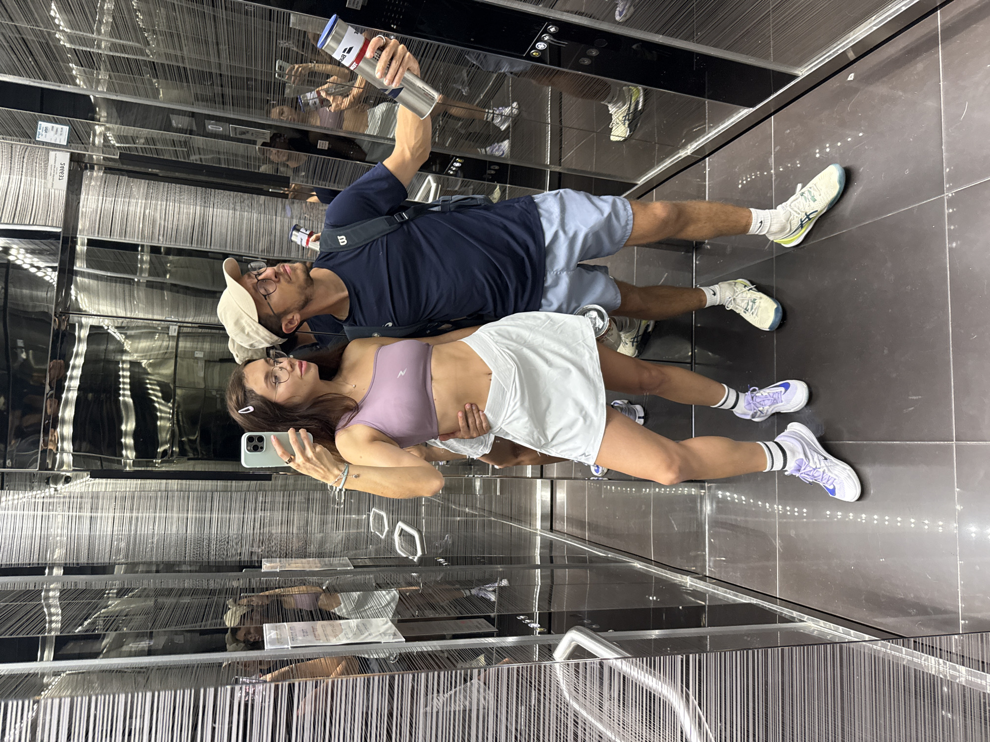 A couple is posing for a selfie in an elevator. The woman is holding a light green smartphone and is wearing a light purple sports bra and a white tennis skirt. The man behind her is wearing a navy blue t-shirt, light blue shorts, a beige baseball cap, and glasses, and he is holding a metallic water bottle. The elevator interior is mirrored and lined with what appear to be vertical silver strands. The floor is dark grey tile. The photo was taken in the city of Tel-Aviv, Israel.