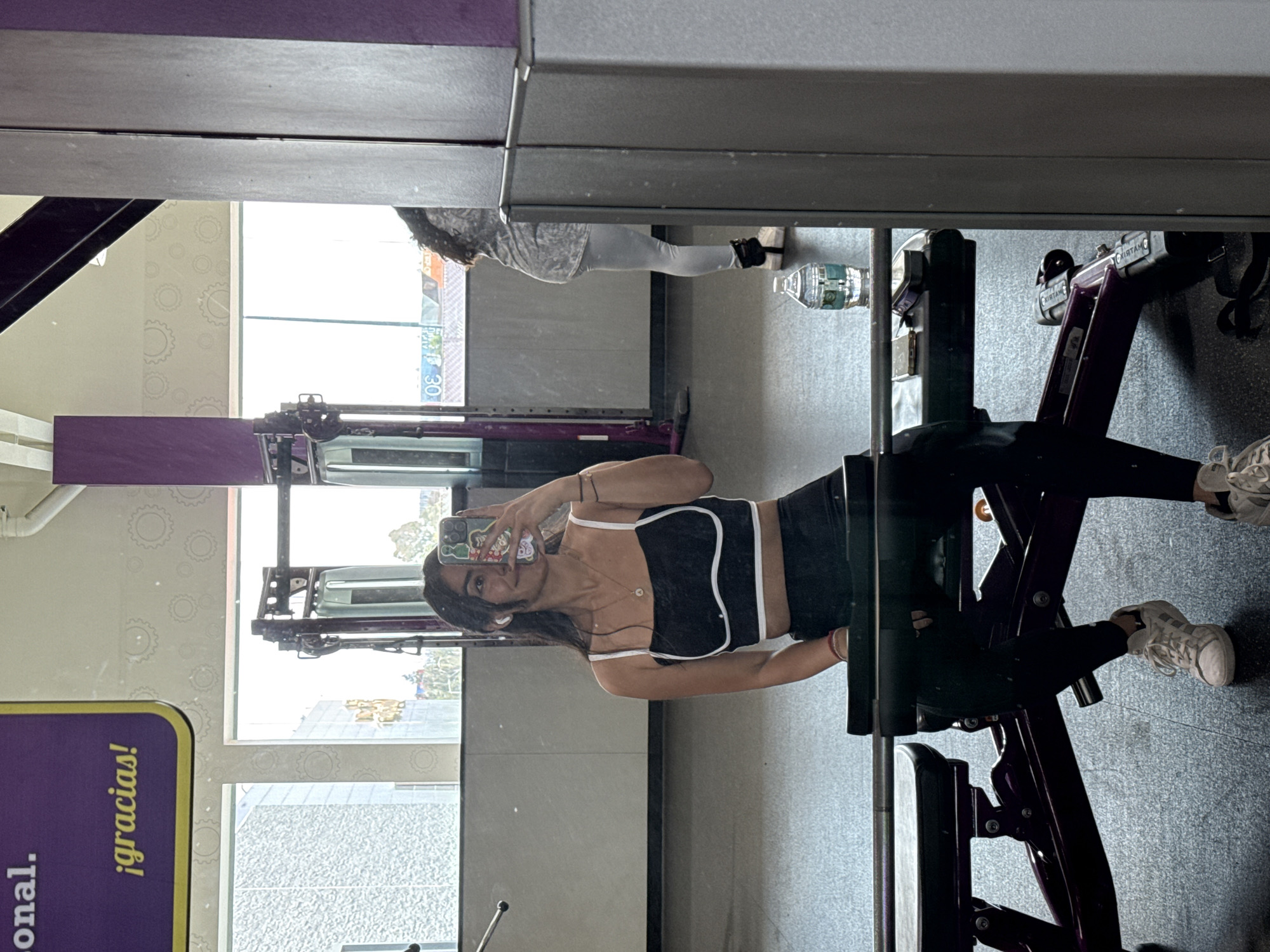 This is a mirror reflection of a young woman taking a selfie inside a gym, likely located in Puebla, Mexico, given the "¡gracias!" sign. The image is oriented vertically (rotated 90 degrees clockwise).
In the foreground, a woman stands facing the mirror, holding a smartphone with a colorful cartoon-stickered case to capture her reflection. She is wearing a black athletic crop top with white trim and thin straps, black leggings, and white sneakers. Her hair is dark and falls over her shoulders. She wears a necklace and small earrings, and a white earbud is visible in her right ear. She has a slight smile.
The setting is a gym, characterized by dark purple and black exercise equipment, including what appears to be part of a leg press machine. The floor is dark gray with a textured pattern. In the background reflection, bright daylight streams through large windows, revealing glimpses of the outside, possibly buildings or an urban environment. Another person, dressed in light-colored athletic wear, is partially visible in the distance, also reflected in the mirror, standing near a plastic water bottle.
Visible text includes "¡gracias!" on a purple sign with yellow lettering on the left side of the frame. Another partially obscured sign or advertisement with the number "30" can be seen through the window reflection. The bright lighting suggests it is daytime.