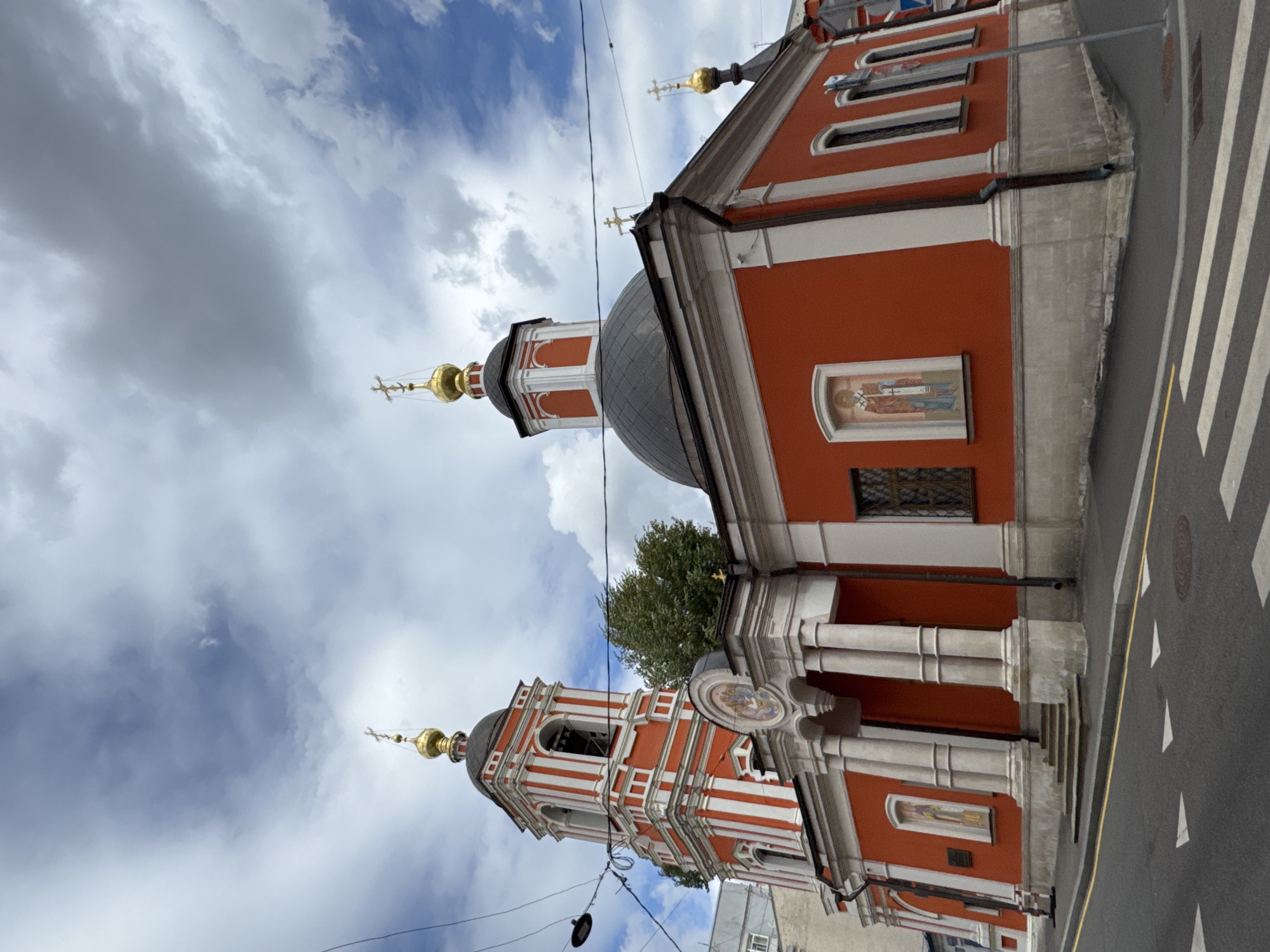 The image shows the Church of St. Nicholas in the Khamovniki district of Moscow, Russia. The church is painted a vibrant orange-red and features white decorative elements. It has a tall bell tower with red and white stripes, topped with a golden dome and cross. The main building has a larger, dark dome, also topped with a cross. There are several windows visible, and ornate religious icons are displayed on the exterior walls. The church is situated on a street with a pedestrian crossing and visible road markings. The sky is partly cloudy with a mix of blue sky and grey clouds.