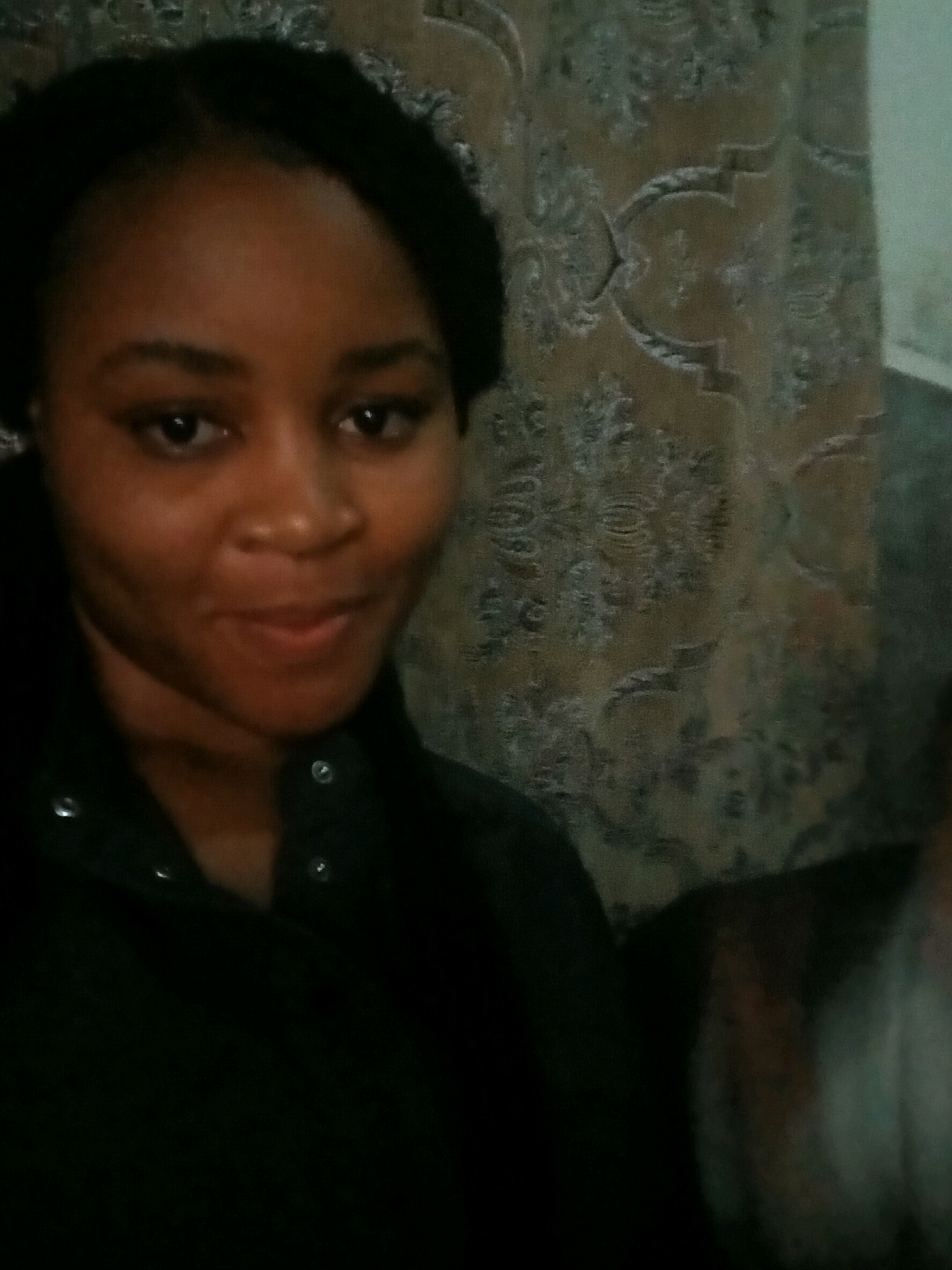 A portrait of a young Black woman is captured in a medium close-up shot, facing the camera with her head slightly turned to her left. Her eyes are dark and appear to be looking forward, and her lips are slightly parted, suggesting a natural, unposed expression. She has dark, textured hair styled in a way that frames her face. She is wearing a dark, collared garment that fastens with buttons, which are visible along the placket. The background consists of a textured fabric, possibly a curtain or upholstery, with an ornate, possibly damask-like pattern in muted tones. The lighting is dim, casting a slight shadow on her right cheek and creating a generally subdued atmosphere. To the far right of the frame, a blurry, lighter-colored element suggests the presence of another person or object, though it is not clearly defined. The image appears to have been taken indoors, possibly in a residential setting, given the domestic nature of the background. There are no discernible city-specific landmarks or context to place the scene within a particular location in Unknown, Unknown.