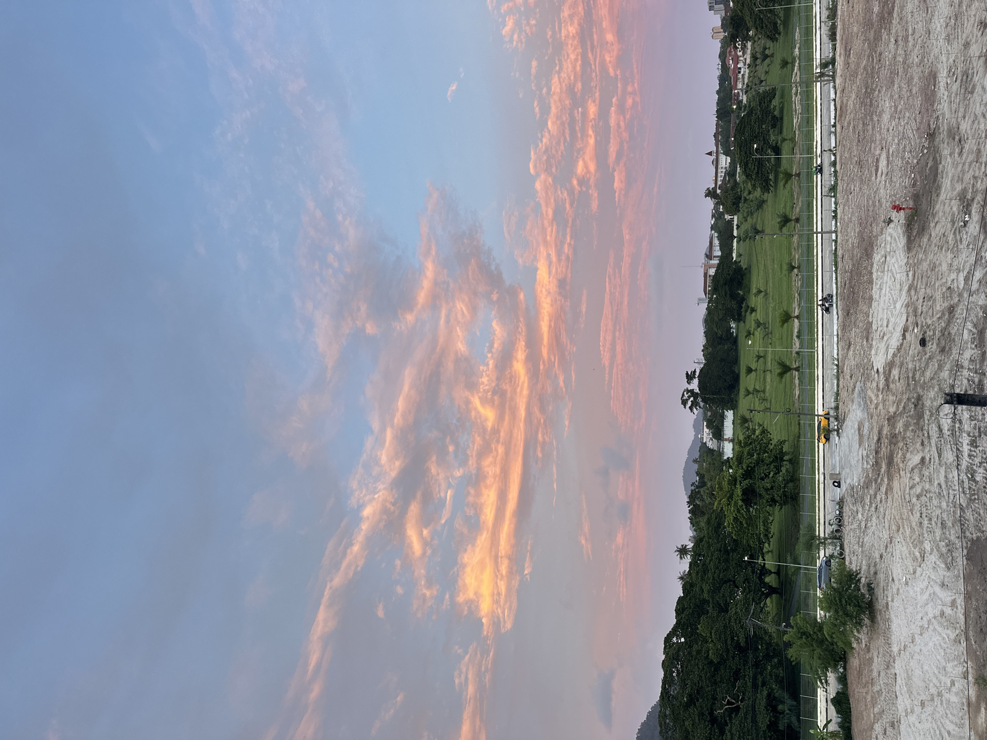 The image depicts a serene twilight scene in Wichit, Thailand. A vibrant sky painted with hues of pink, orange, and blue dominates the upper half of the frame, with wispy clouds catching the last rays of sunlight. Below the sky, a lush green landscape unfolds, dotted with scattered palm trees and larger deciduous trees. In the mid-ground, a road runs horizontally across the image, with a few vehicles, including motorcycles and bicycles, present. A yellow car is parked on the side of the road. The background features a cityscape with several buildings and hills. The foreground consists of a construction site with dirt and exposed ground, suggesting ongoing development. The overall atmosphere is peaceful and tranquil, capturing the beauty of a sunset over a developing urban area.