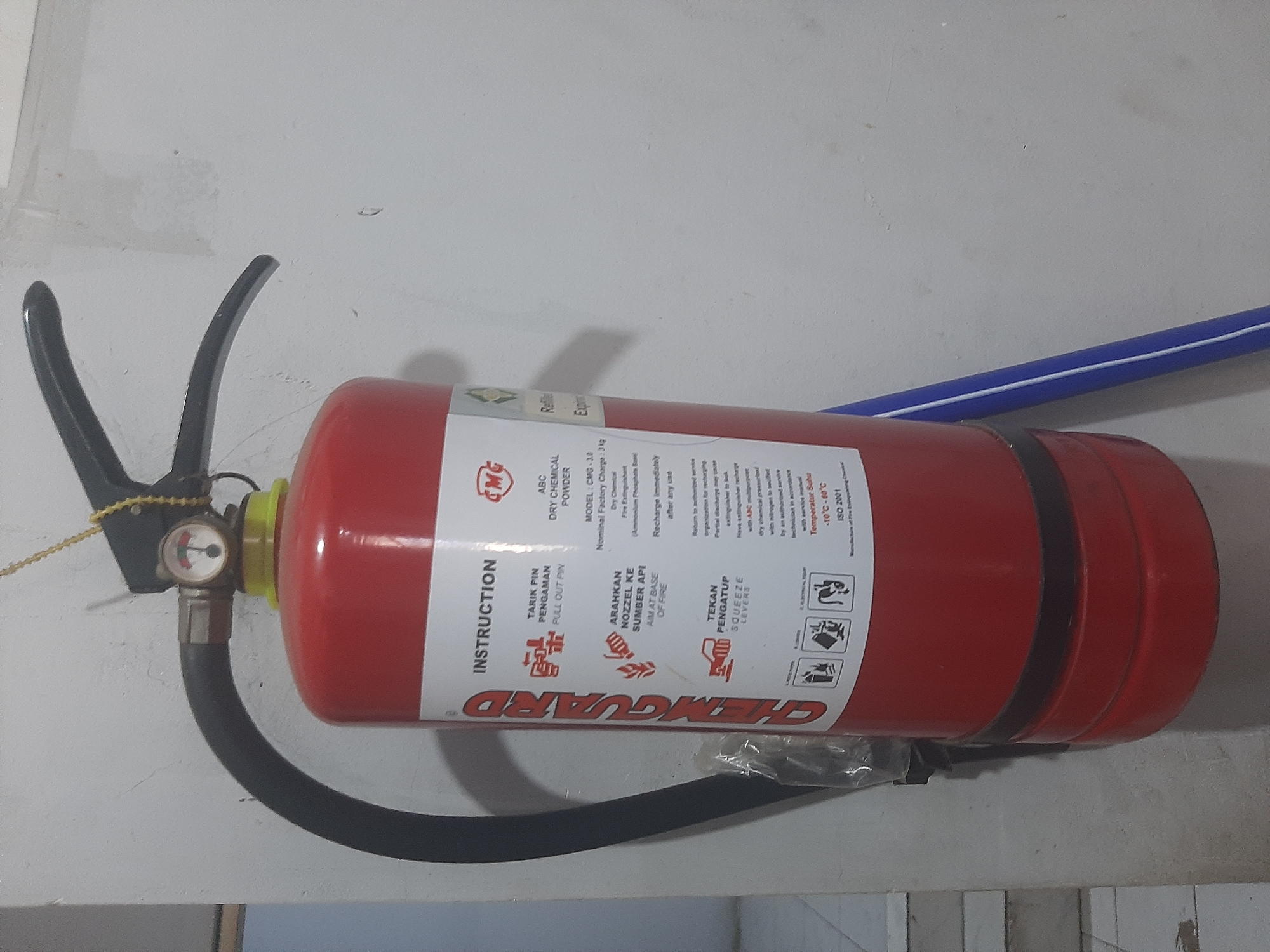 An ABC dry chemical powder fire extinguisher, identified as CHEMGUARD, is mounted on a light-colored wall. The extinguisher is red with a black hose and handle. A pressure gauge is visible at the top of the cylinder. The label provides instructions for use in Indonesian and English, including "PULL OUT PIN," "AIM AT BASE OF FIRE," and "SQUEEZE LEVERS." It also lists the model as CMG-3.0, nominal factory charge as 3 kg, and the operating temperature as -10°C to 60°C. The extinguisher is positioned to the left of a blue and white striped object, possibly a pipe or pole, leaning against the wall. The environment appears to be indoors, with a floor visible in the lower left corner. No people are present. The overall scene is utilitarian, suggesting a safety or emergency preparedness context. The location is West Jakarta, Indonesia.